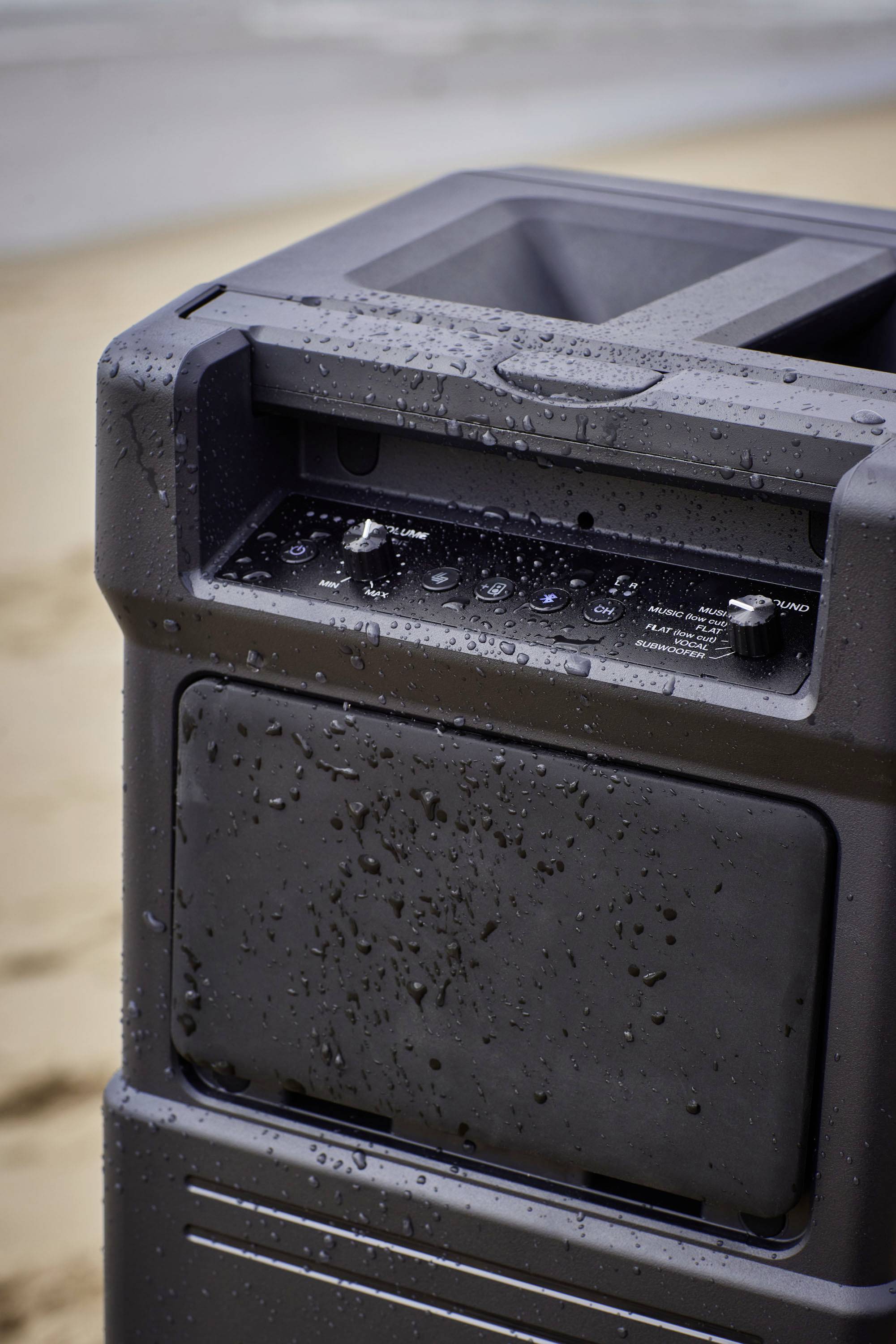 Ein nasser, tragbarer Lautsprecher am Strand zeigt Regler und Knöpfe an der Oberseite. Hintergrund unscharfer Sand und Meer.