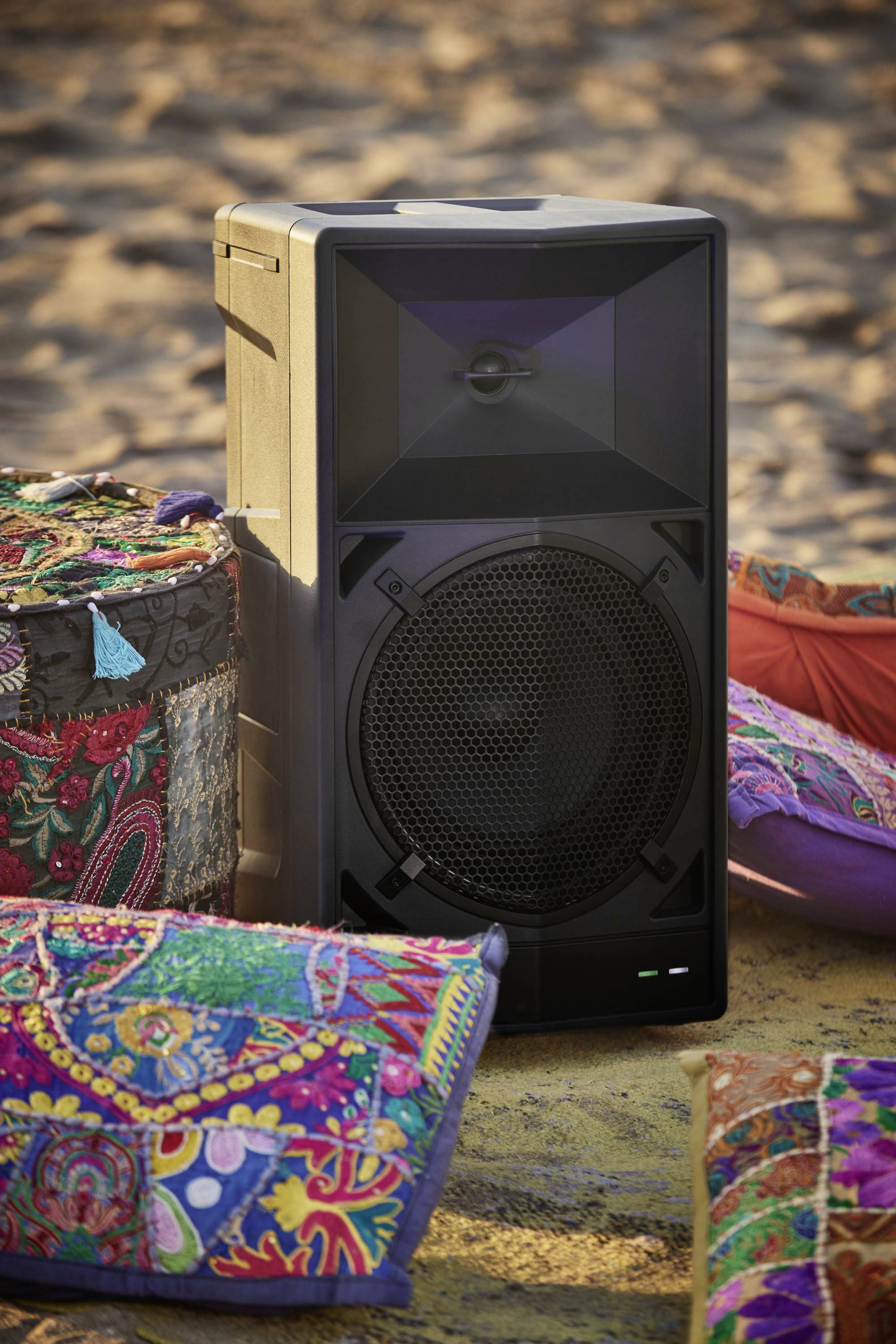 Boxenlautsprecher im Freien auf Sand platziert, umgeben von bunten Kissen; geeignet für Musikgenuss an einem sonnigen Tag.