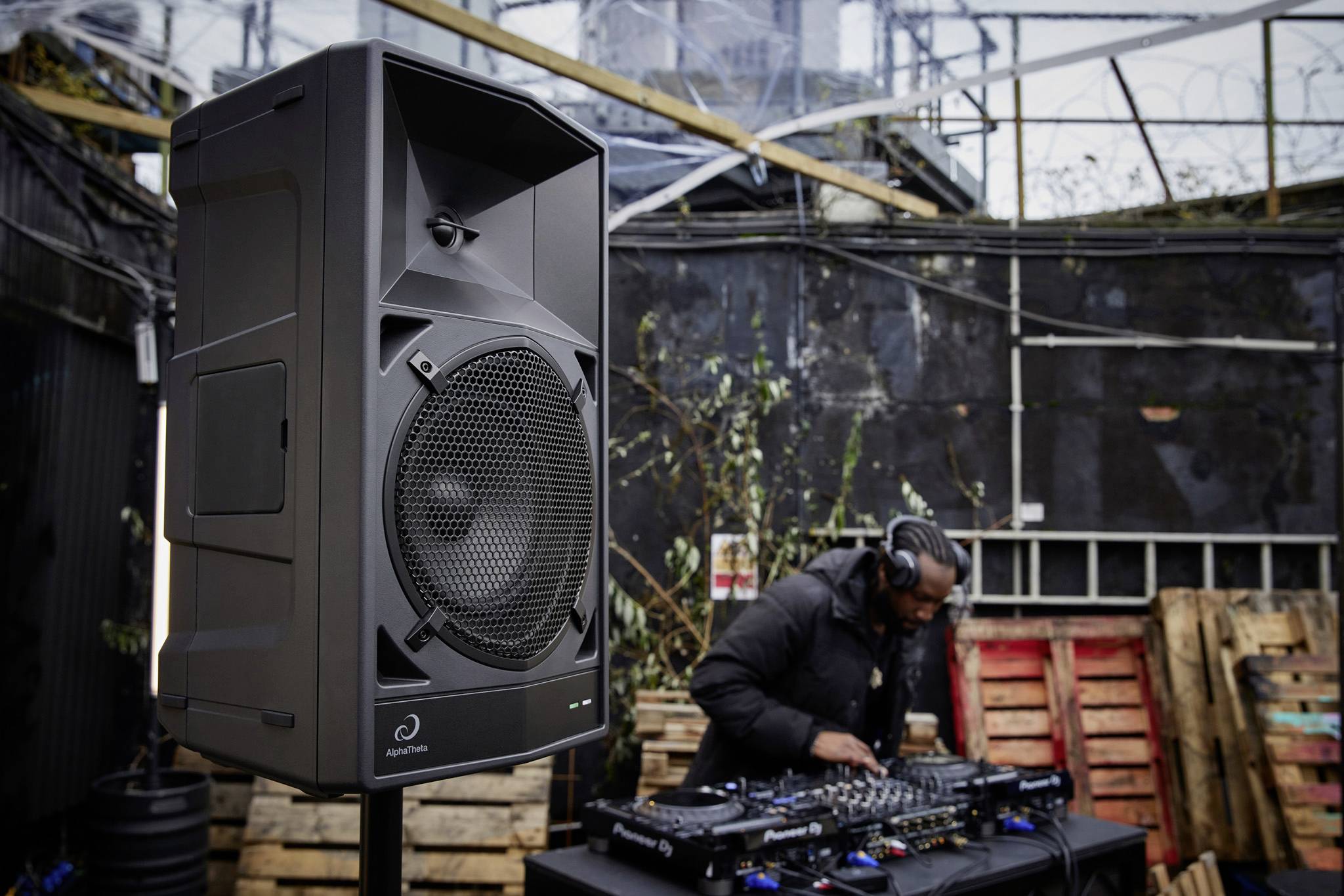 Eine Person legt Musik auf DJ-Equipment auf, während im Vordergrund ein großer Lautsprecher zu sehen ist. Im Hintergrund ist eine industrielle Umgebung.