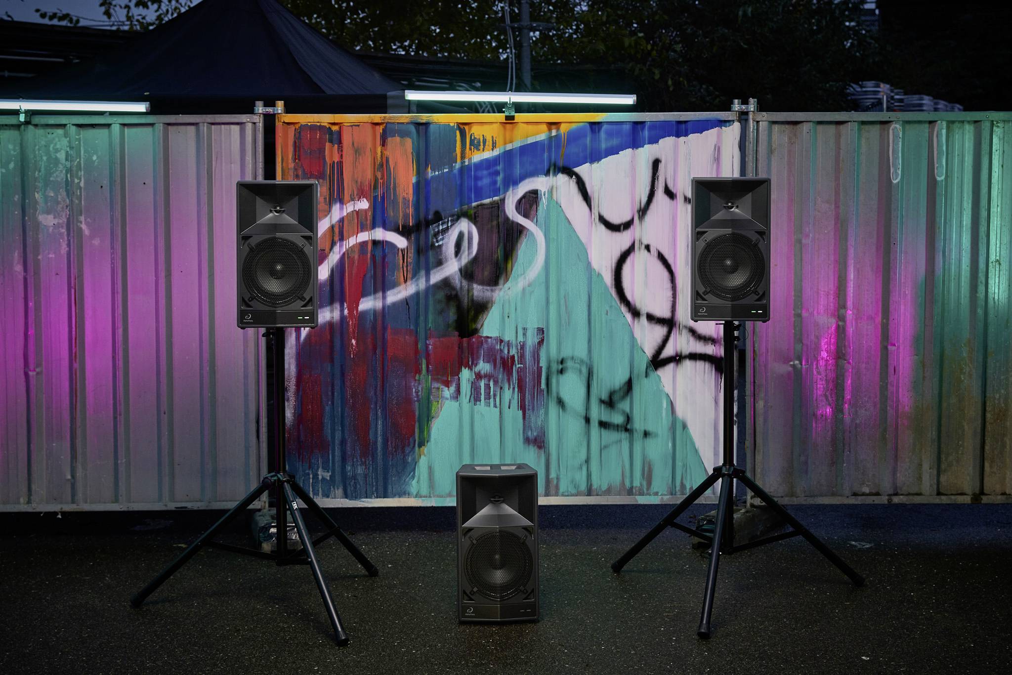 Drei Lautsprecher stehen vor einer bunt bemalten Wand mit Graffiti. Die Wand ist mit farbigen Lichtern beleuchtet, die Stimmung erzeugen.