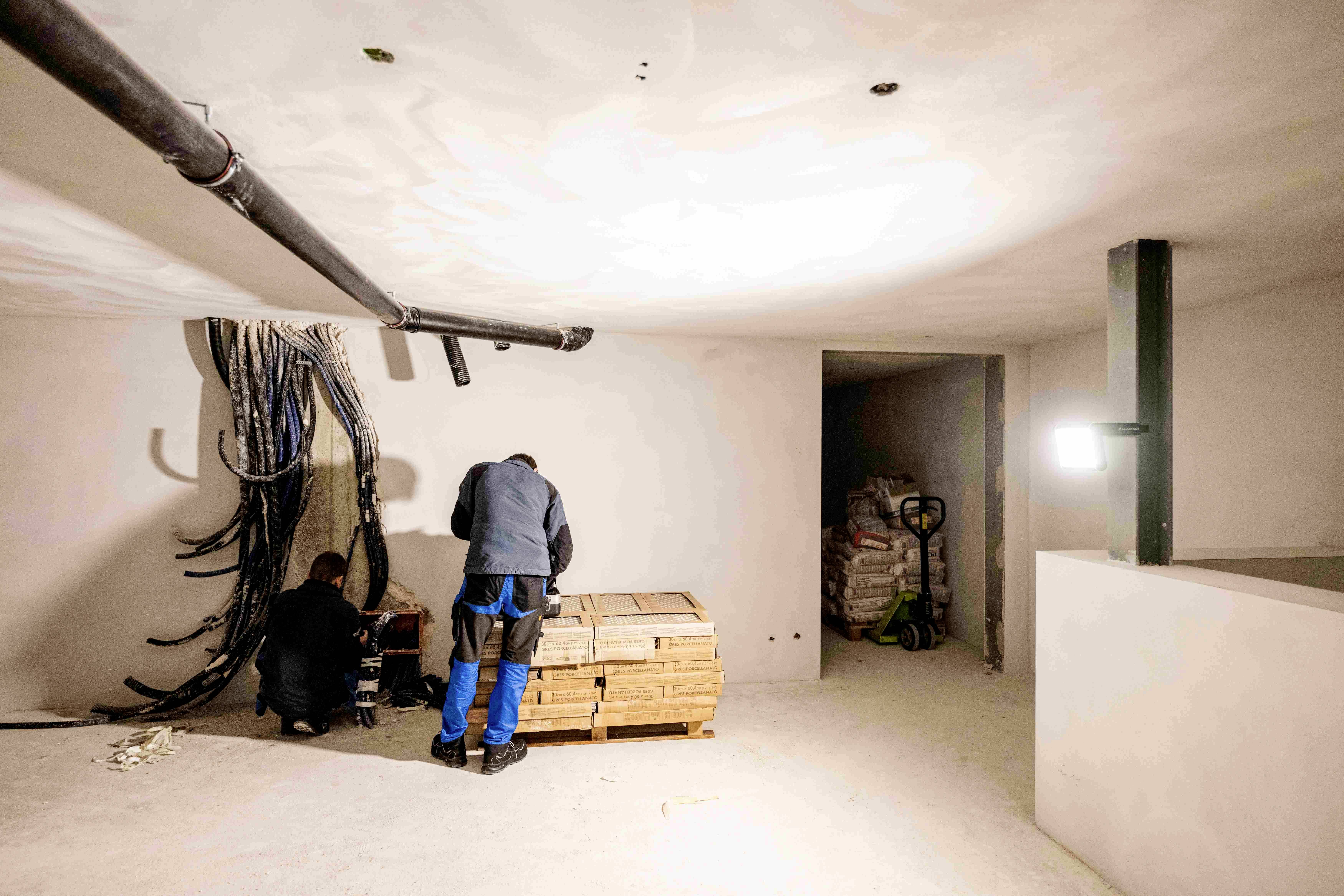 Zwei Personen arbeiten in einem hellen Raum an Kabeln an der Wand. Im Hintergrund liegen Baumaterialien. Raum wirkt unfertig.