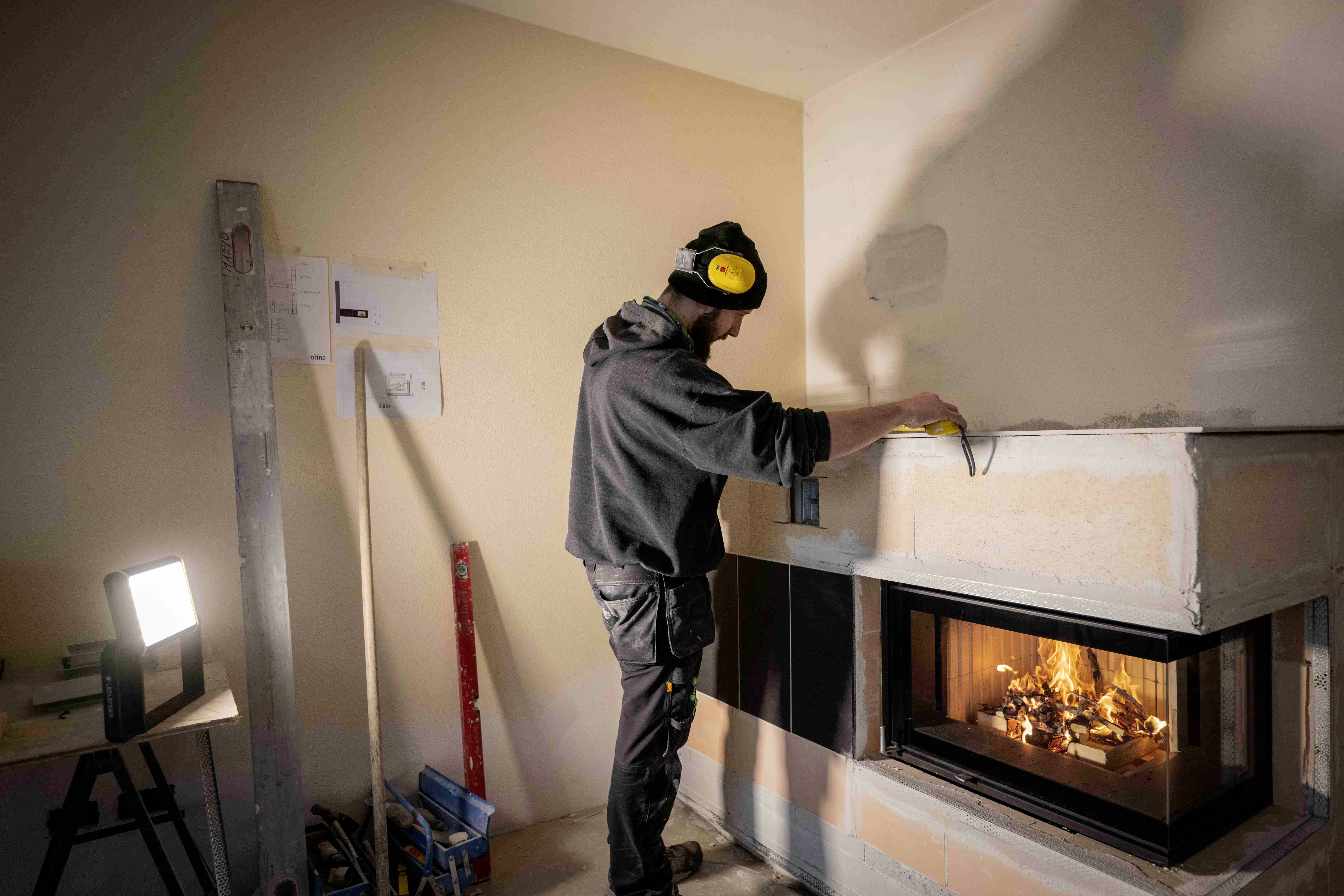 Eine Person, die in einem Raum einen Kamin installiert. Schutzkleidung und Werkzeuge sind sichtbar, während das Feuer im Kamin brennt.