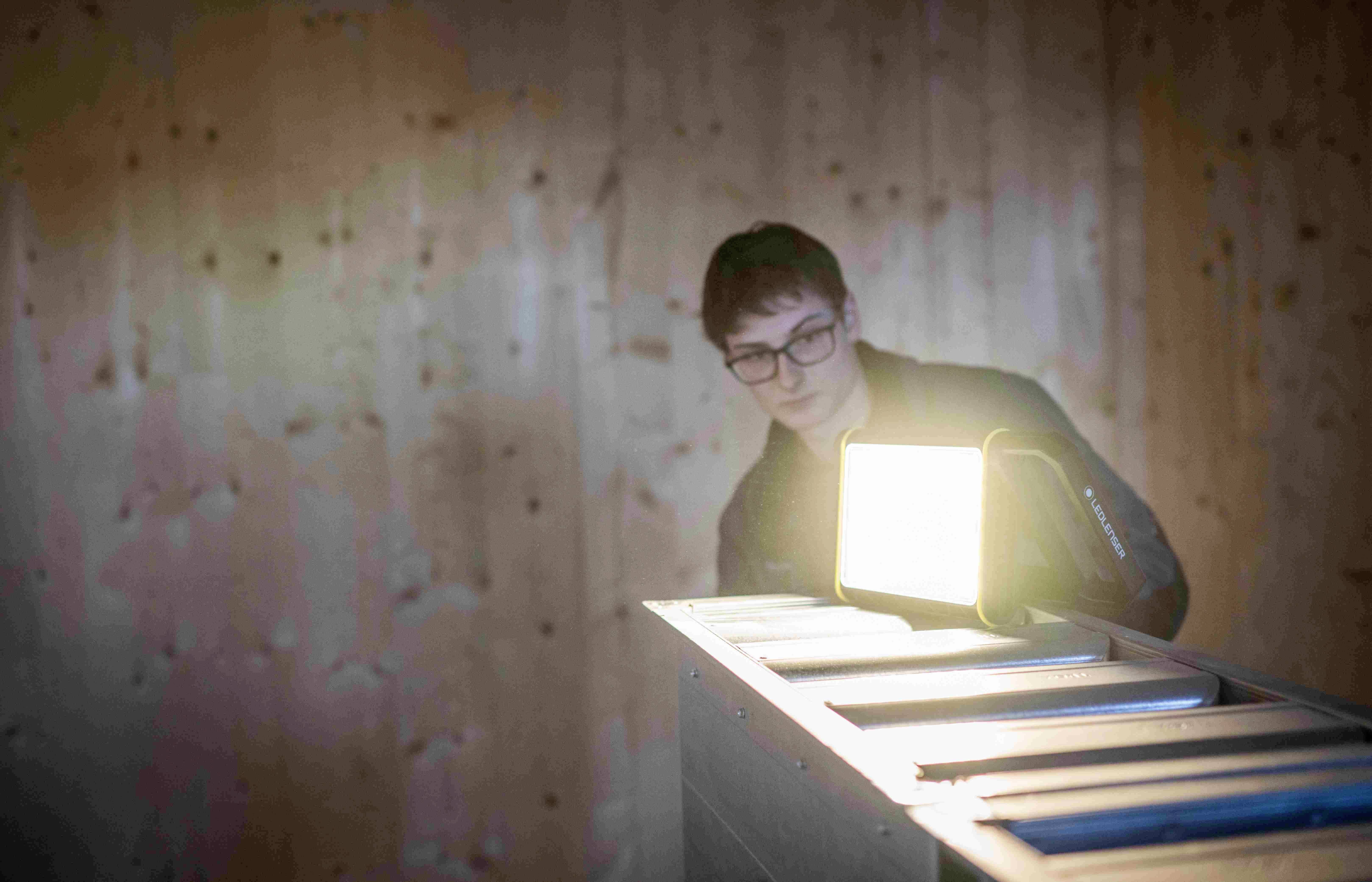 Eine Person mit Brille, hinter einem leuchtenden Licht, steht vor einer Holzwand in einem schummrig beleuchteten Raum.