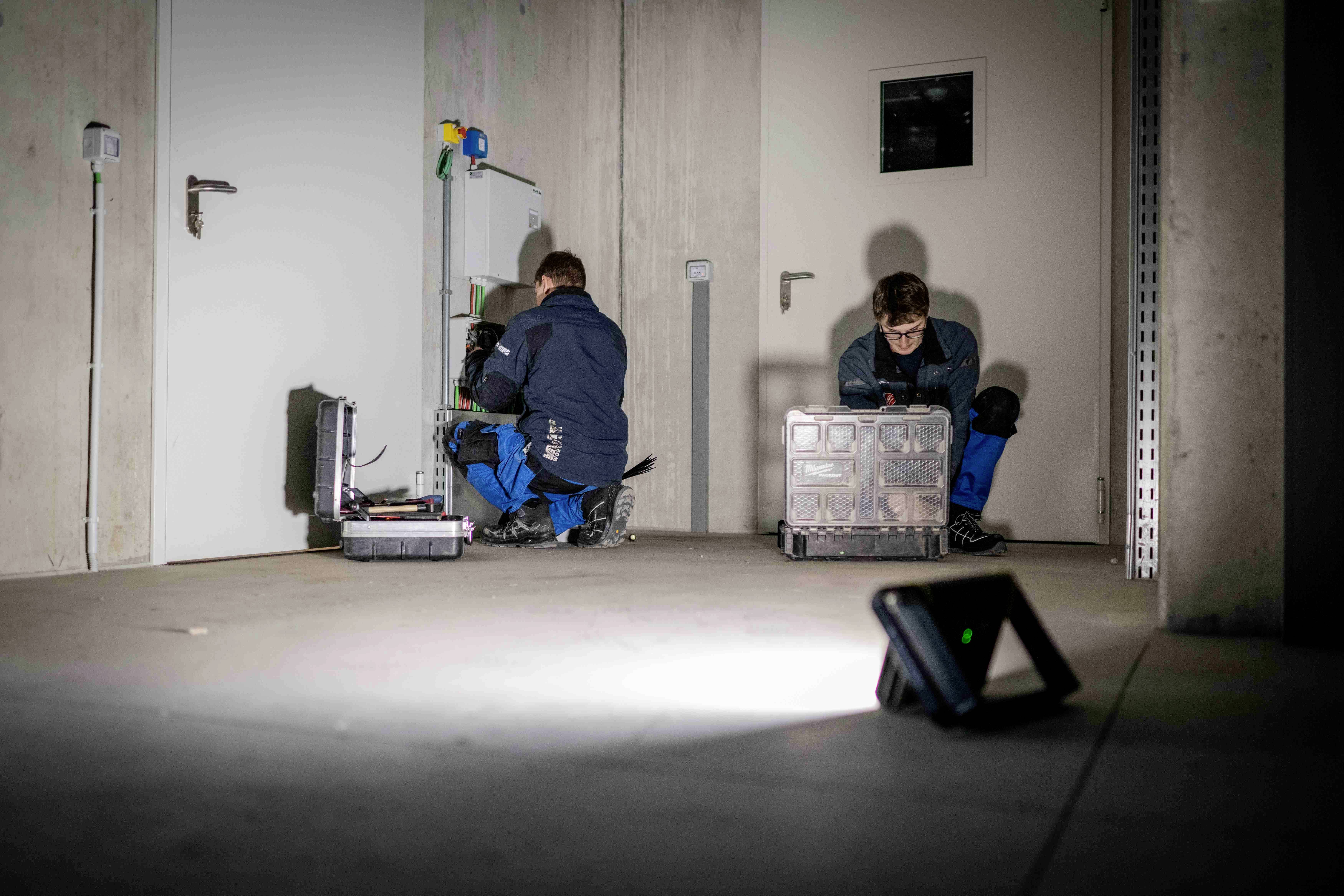Zwei Personen in Arbeitskleidung arbeiten in einem Technikraum. Eine Person überprüft elektrisches Gerät, während die andere Kisten sortiert.