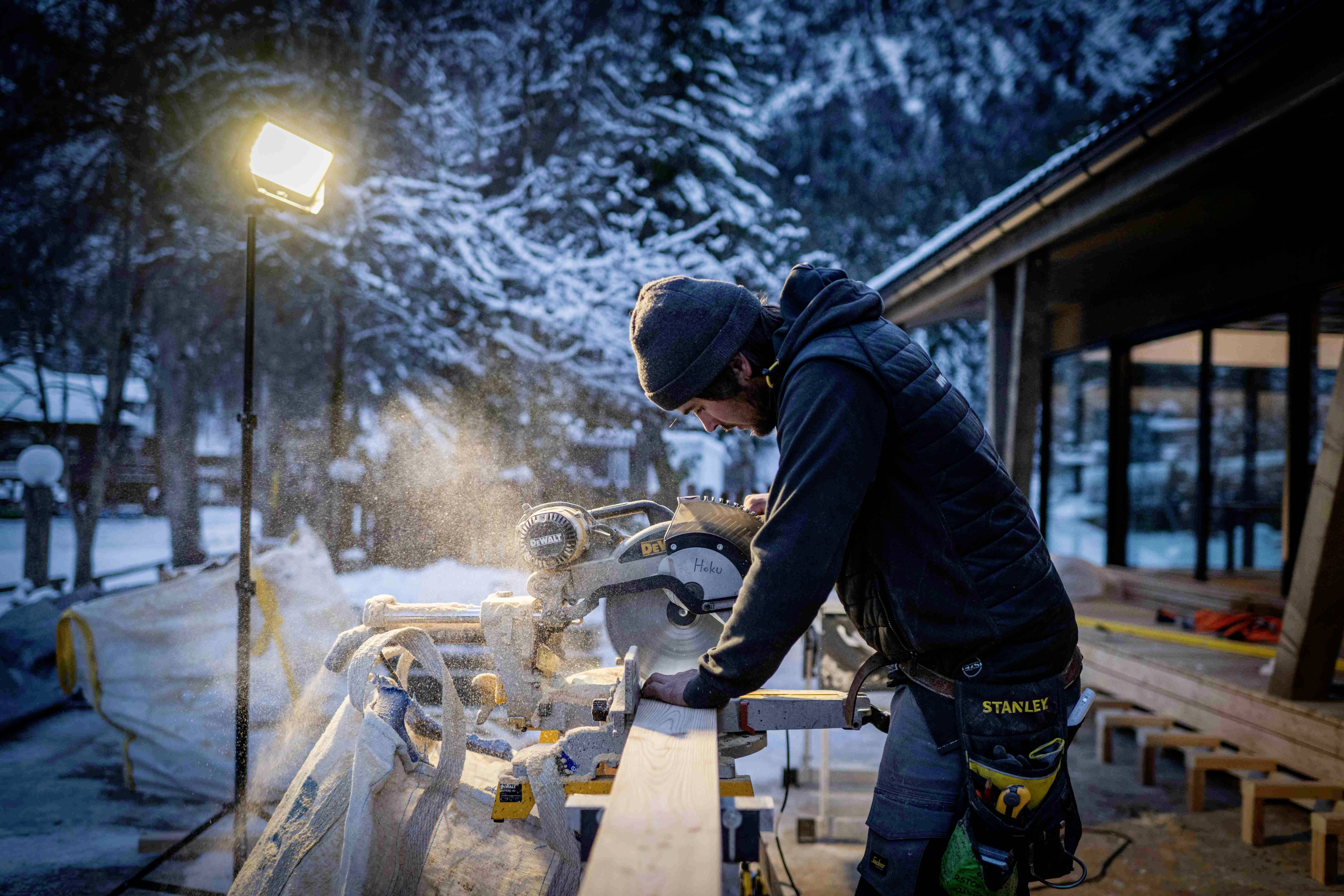 Ein Handwerker sägt ein Holzbrett mit einer Kreissäge im Freien. Es ist Winter, Schnee bedeckt den Boden, und eine Lampe beleuchtet die Szene.