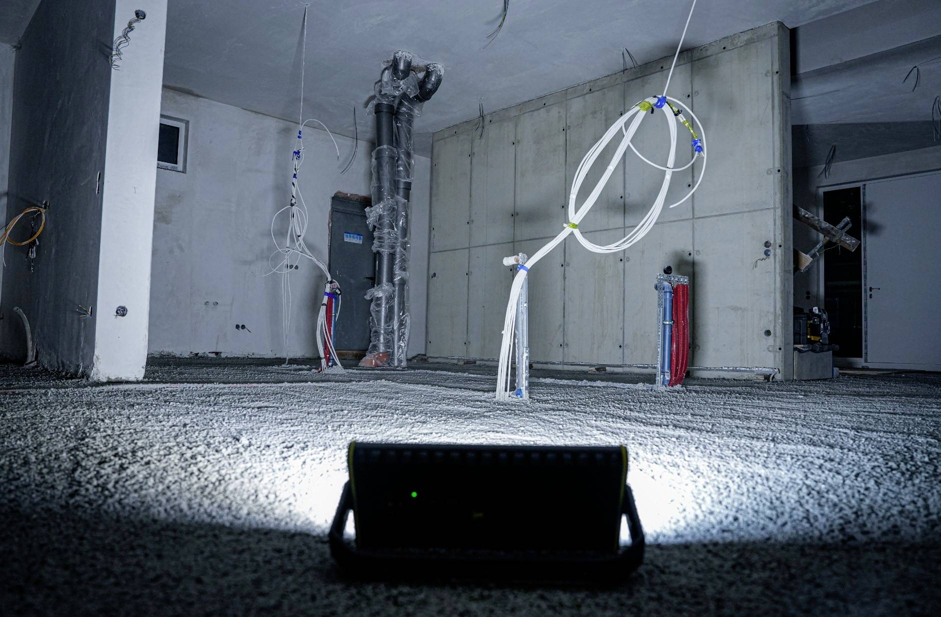 Kellerraum mit Betonestrich und freiliegenden Kabeln, beleuchtet von einer Lampe am Boden. Wände unverputzt, Baustellenatmosphäre.