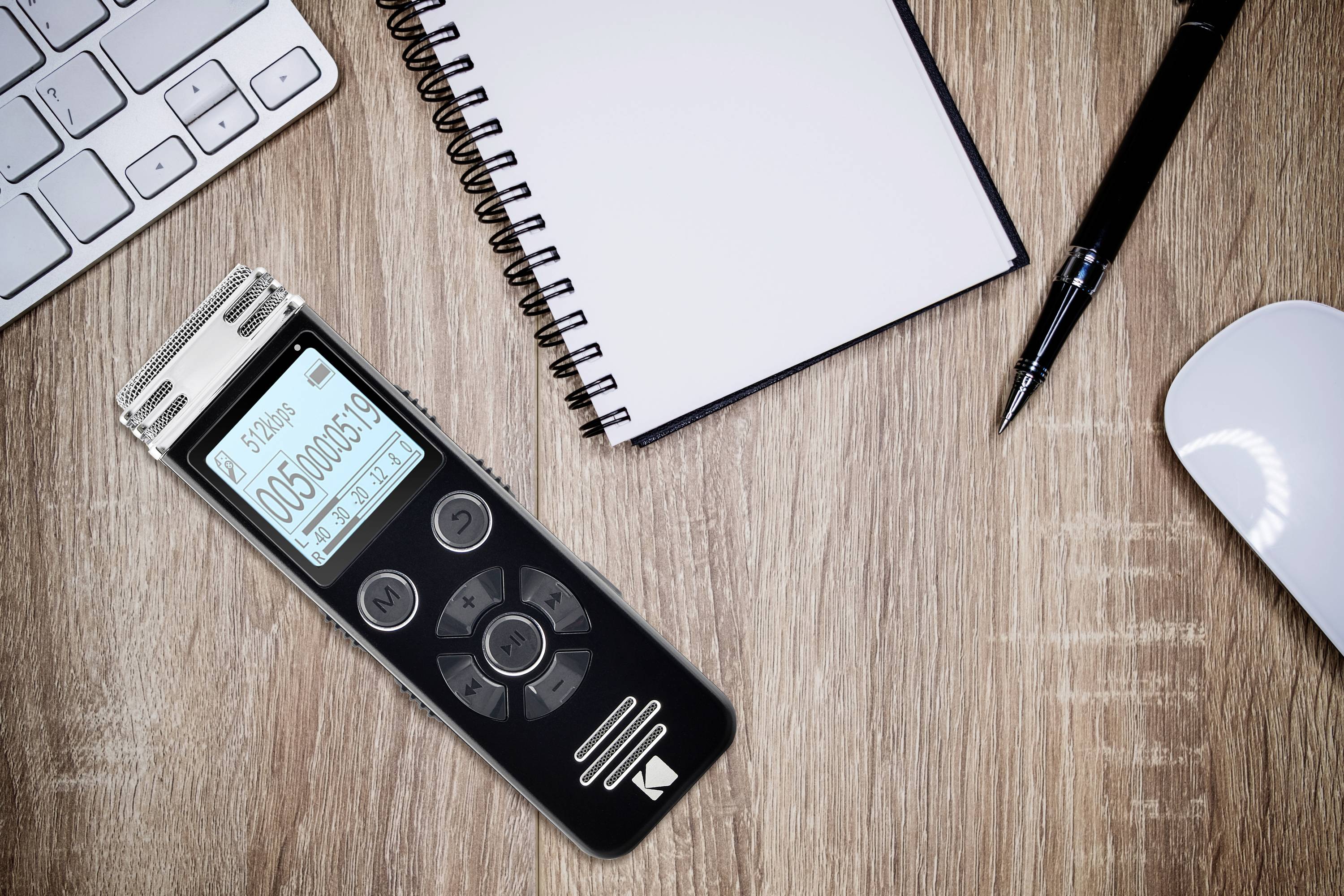 Ein digitaler Audiorecorder liegt auf einem Holztisch neben einem Notizblock, einem Stift, einer Tastatur und einer Maus.