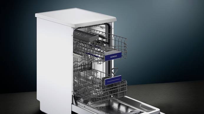 Ein Standgeschirrspüler von Siemens steht in einem leeren Raum mit schwarzem Boden und dunkelblauer Wand. Die Tür steht komplett offen und beide Schubladen sind herausgezogen. In der unteren befindet sich ein Besteckkorb. Es befindet sich kein Geschirr in der Maschine.