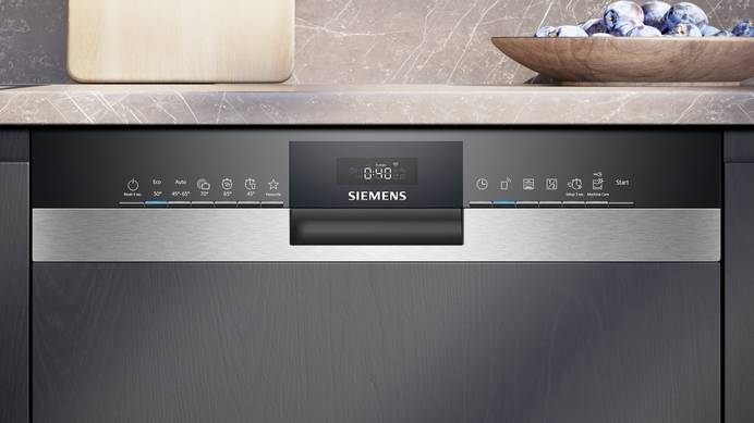 Ein teilintegrierter Einbaugeschirrspüler von Siemens ist in eine dunkle Küchenzeile eingebaut. Wie anhand der Bedienelemente zu erkennen ist, läuft die Maschine im Eco-Modus. Das Display zeigt an, dass der Spülgang noch 40 Minuten lang läuft. Auf der Arbeitsfläche über dem Geschirrspüler stehen ein Schneidbrett und ein Teller mit Pflaumen.