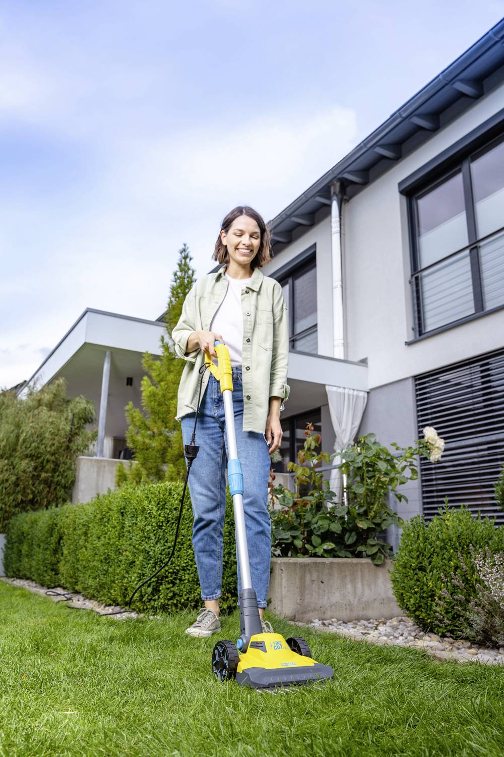 Eine Frau mäht den Rasen mit einem elektrischen Rasenmäher vor einem modernen Haus. Es ist sonnig und der Garten ist gepflegt.