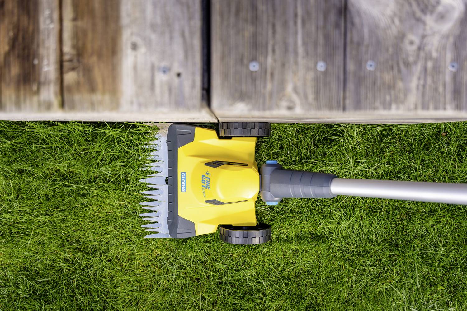 Ein gelb-schwarzer Rasentrimmer mit Rädern schneidet Gras entlang einer Holzterrasse. Das Gerät ist auf grünem Rasen platziert.