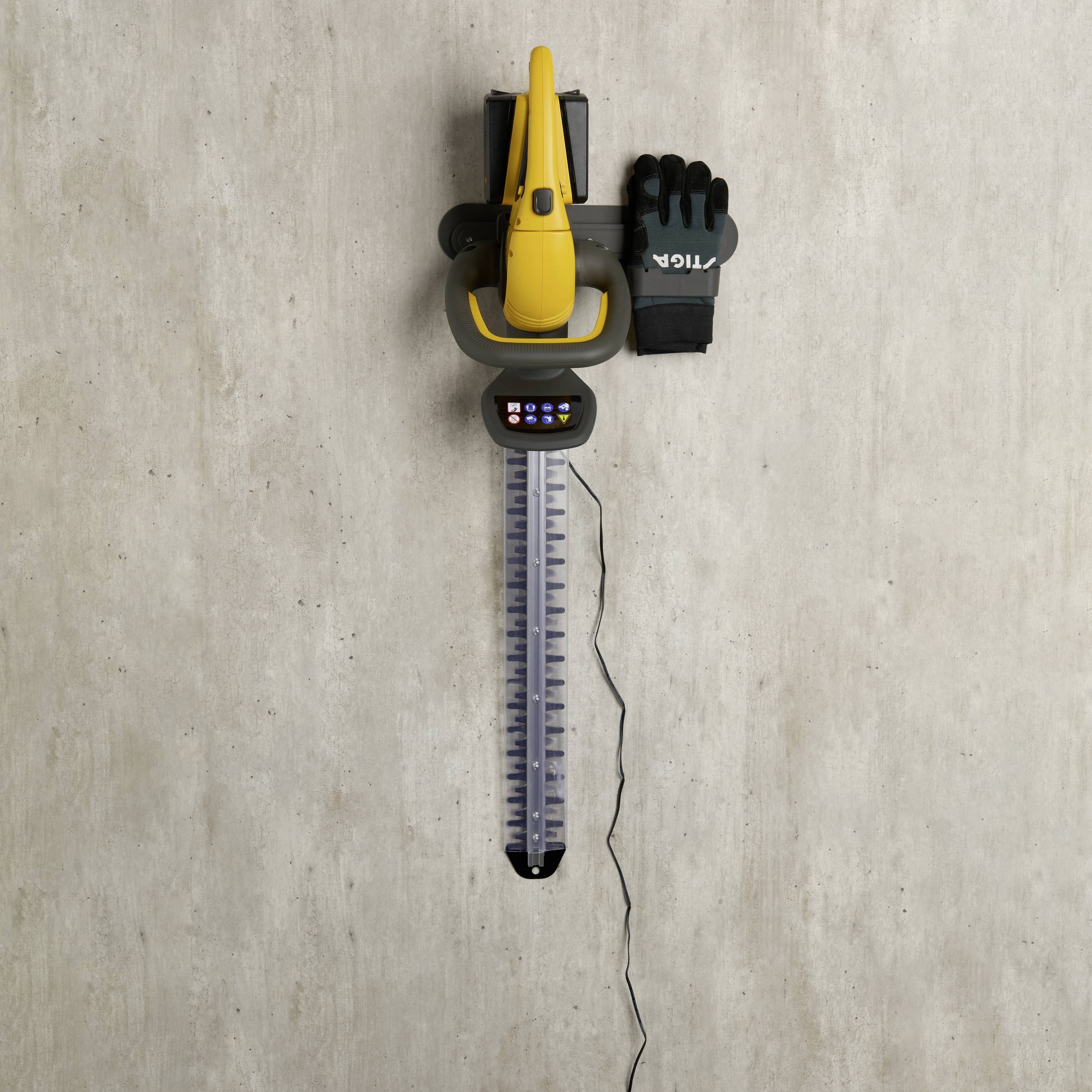 Elektrische Heckenschere hängt an einer grauen Wand. Daneben ein schwarzer Handschuh.