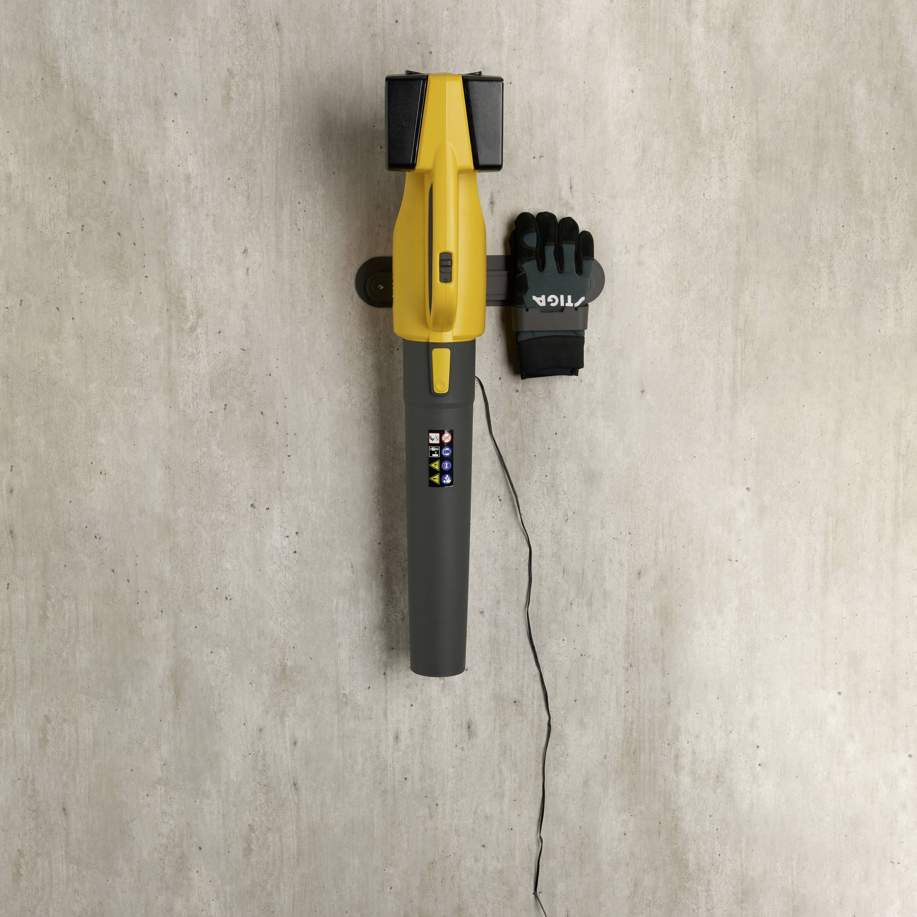 Ein gelber Handstaubsauger hängt an einer Wand, daneben ein schwarzer Handschuh mit 'TIGA'-Logo. Ein Kabel hängt herunter.