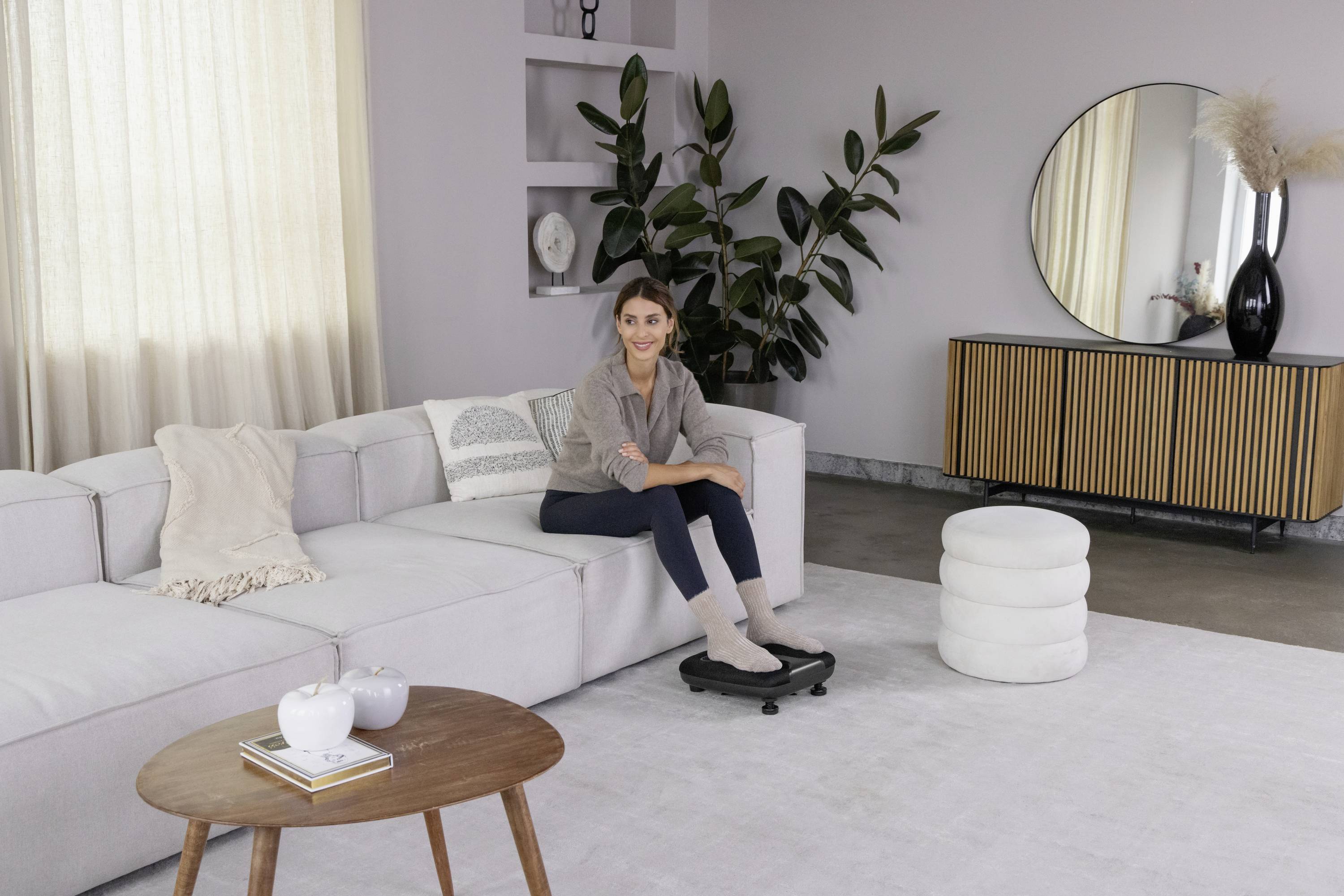 Die Frau sitzt auf einem Sofa in einem modernen Wohnzimmer mit Pflanzen und runden Möbeln. Sie lächelt und schaut zur Kamera.