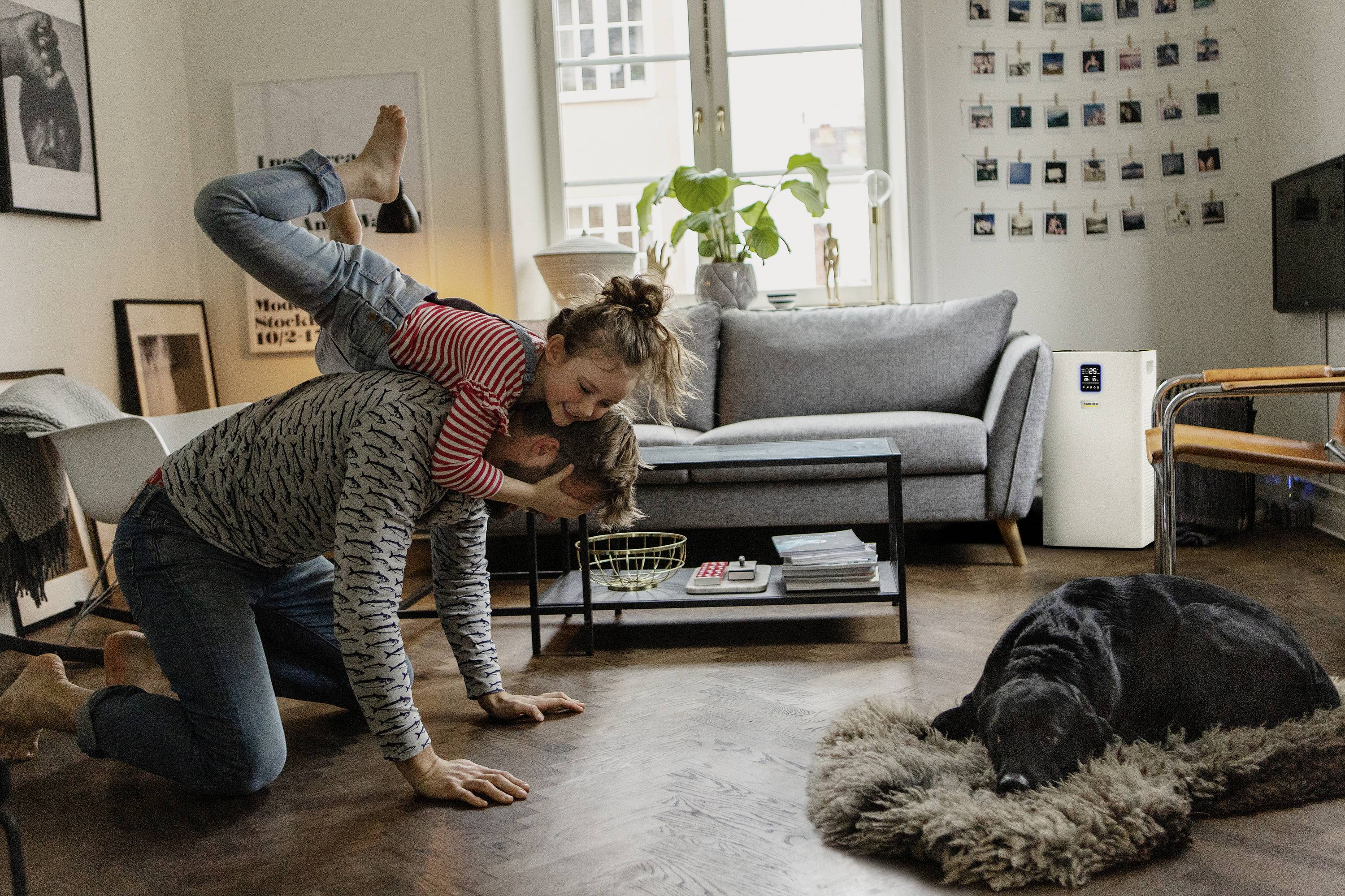 Ein Mann spielt mit einem Mädchen auf seinem Rücken in einem Wohnzimmer. Ein Hund schläft auf einem Teppich. Gemütliche, familiäre Szene.
