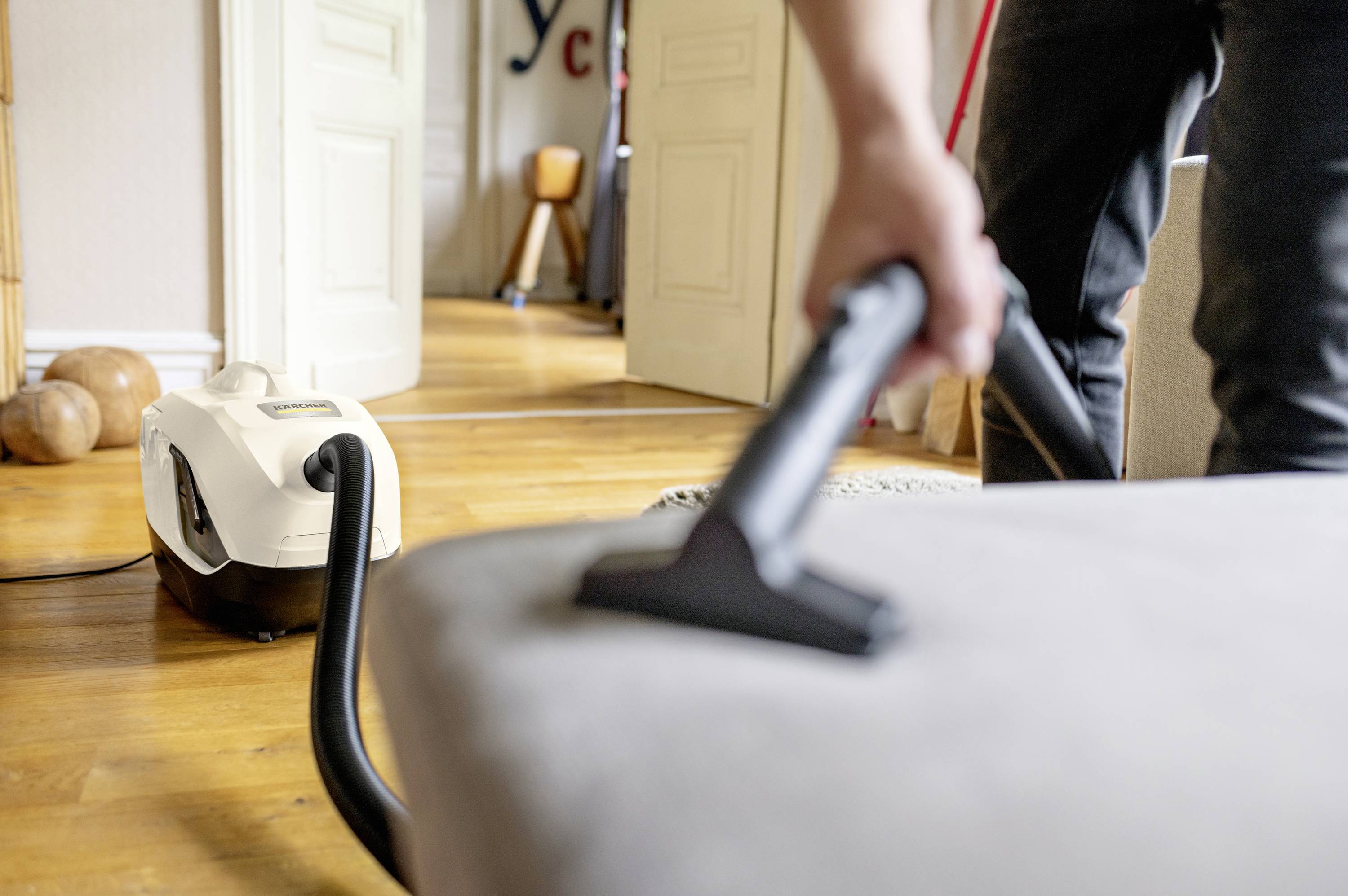 Eine Person saugt mit einem Staubsauger ein Sofa ab. Im Hintergrund sind ein Licht durchflutetes Zimmer und geöffnete Türen zu sehen.