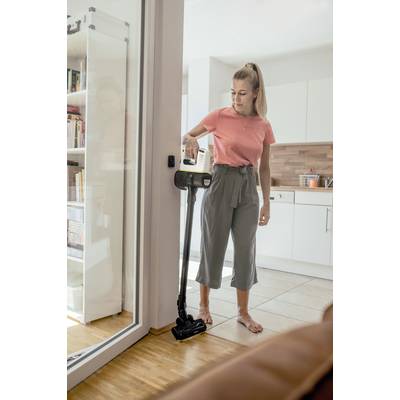 Eine Frau in einem Wohnzimmer saugt den Boden mit einem kabellosen Staubsauger. Im Hintergrund ist eine Küche zu sehen.