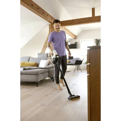 Mann in lila T-Shirt reinigt lächelnd einen hellen Holzboden mit einem kabellosen Staubsauger in einem modernen Wohnzimmer.