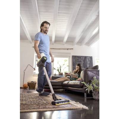 Ein Mann saugt den Teppich in einem modern eingerichteten Wohnzimmer, während eine Frau entspannt auf dem Sofa sitzt.