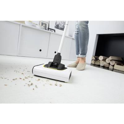 Eine Person in Jeans und beigen Schuhen saugt Krümel vom weißen Teppich in einem Wohnzimmer. Im Hintergrund sind Holzscheite zu sehen.