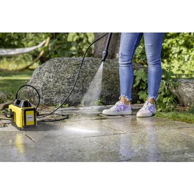 'Person reinigt Steinplatten im Garten mit Hochdruckreiniger. Wasserstrahl entfernt Schmutz, Gras und Bäume im Hintergrund.'