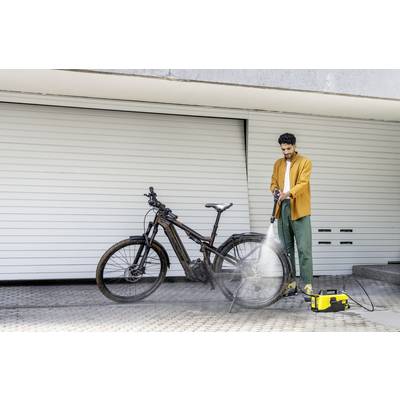 Ein Mann reinigt ein schmutziges Fahrrad mit einem Hochdruckreiniger vor einer geschlossenen Garagentür. Er trägt eine orange Jacke.