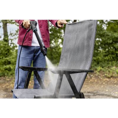Eine Person reinigt einen grauen Gartenstuhl mit einem Hochdruckreiniger im Freien, im Hintergrund sind Bäume zu sehen.