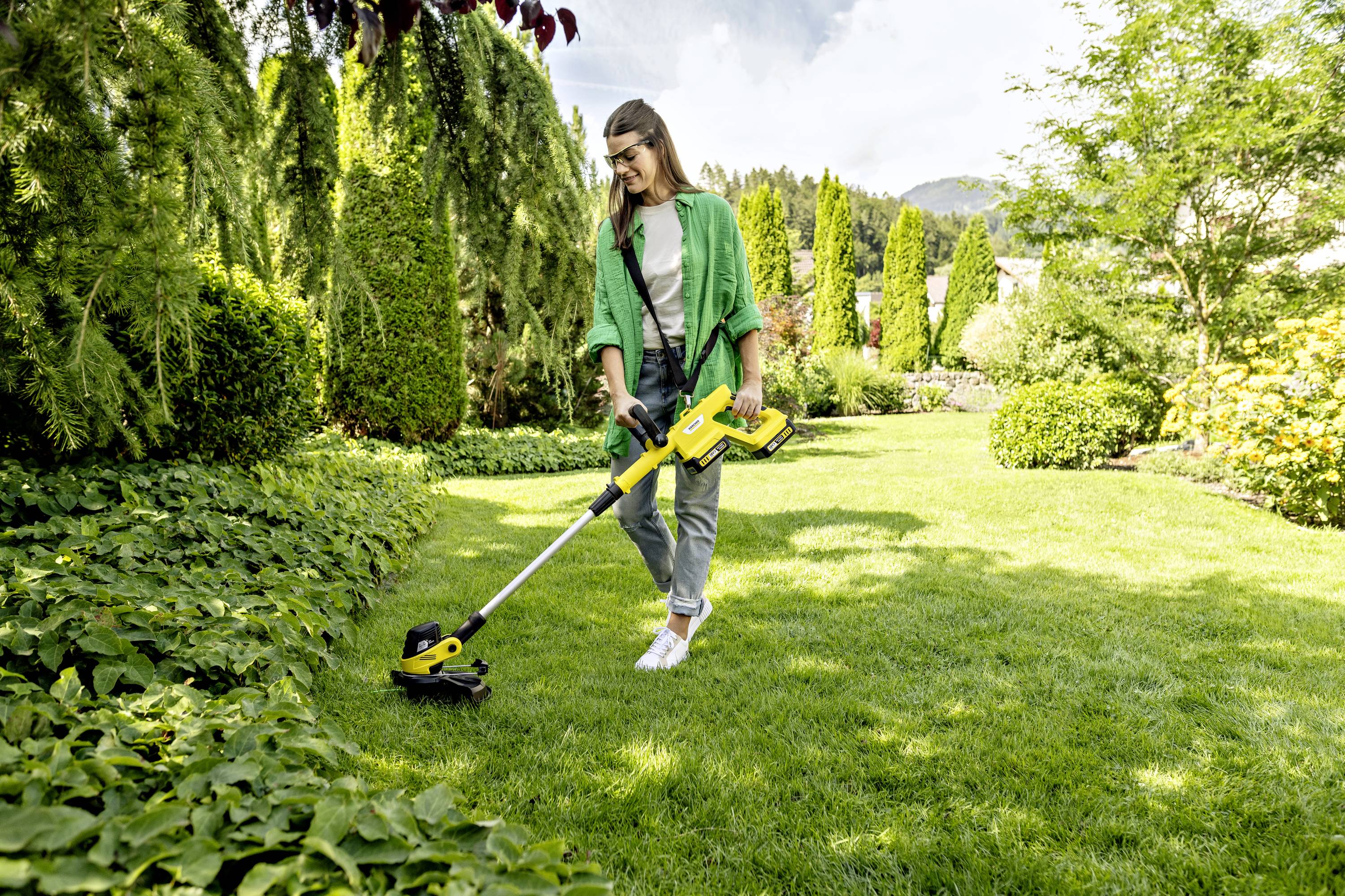 Eine Person mäht mit einem Trimmer den Rasen in einem gepflegten Garten, umgeben von Bäumen und Büschen an einem sonnigen Tag.