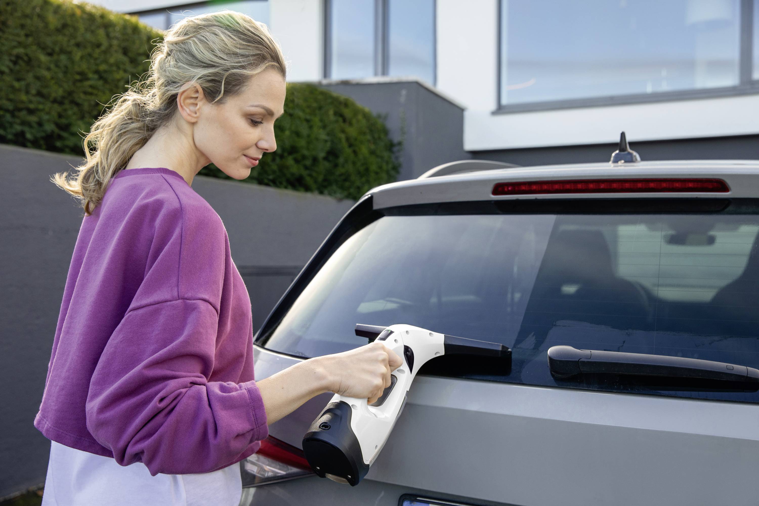 Eine Person reinigt das Heckfenster eines Autos mit einem Fenstersauger. Im Hintergrund sind ein Haus und Büsche zu sehen.