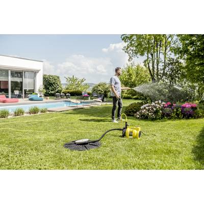Ein Mann bewässert mit einem Gartenschlauch Pflanzen in einem Garten neben einem Pool. Im Hintergrund ist ein modernes Haus zu sehen.