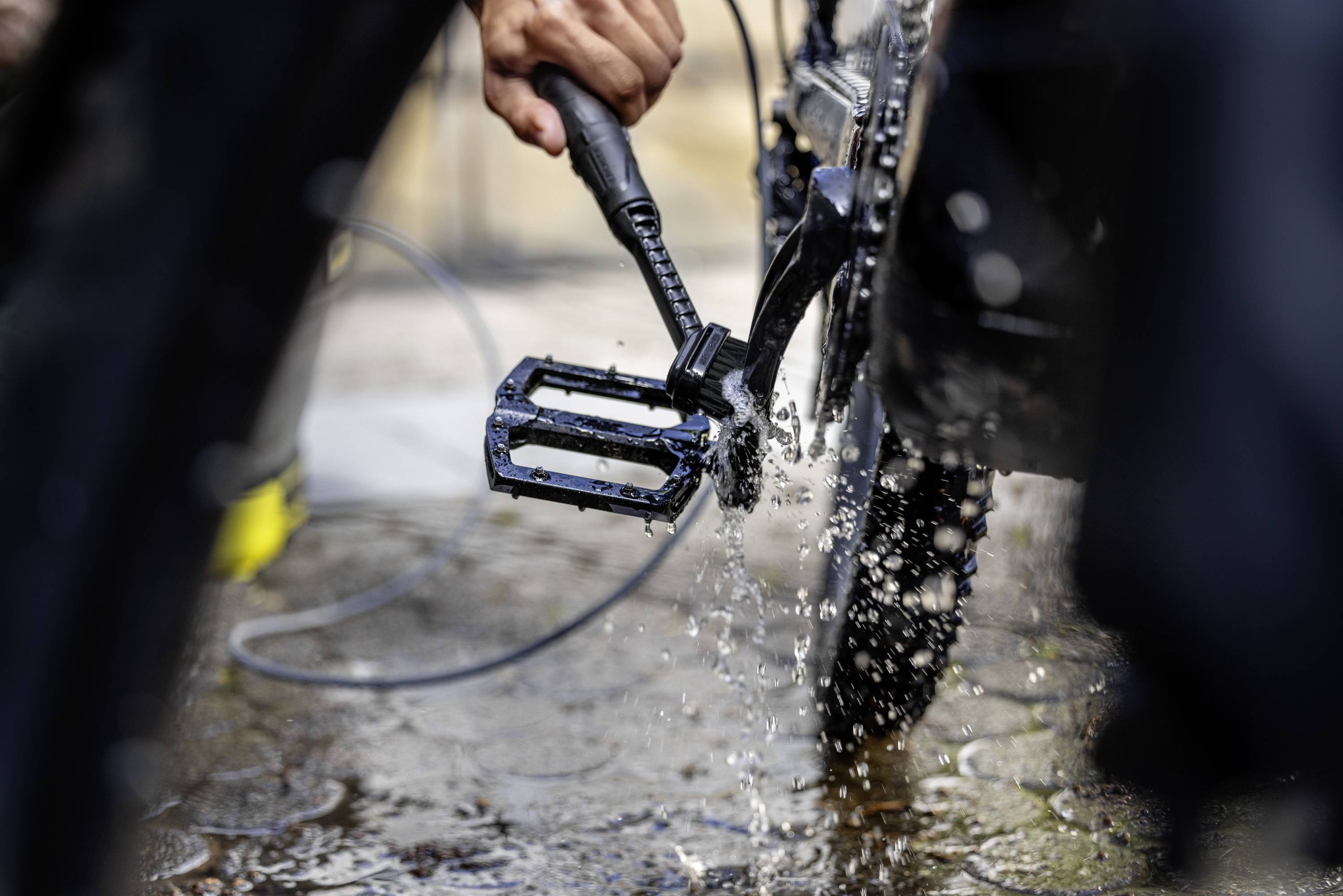 Person reinigt ein Fahrradpedal mit einem Wasserstrahl. Wassertropfen spritzen in die Luft, während das Pedal gründlich sauber wird.