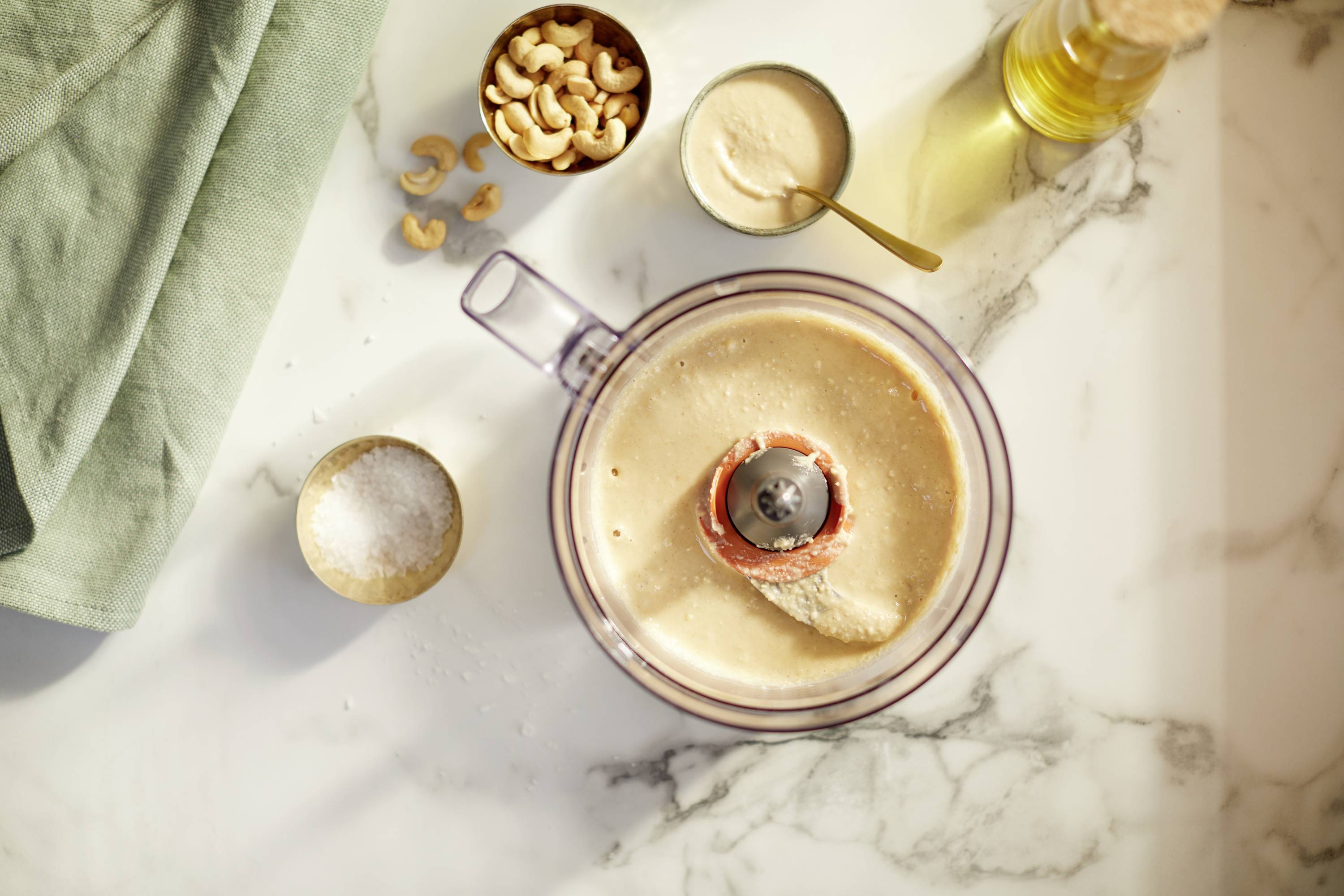Ein Mixer mit Cashewcreme auf einer Marmortheke. Daneben Cashewnüsse, Salz und ein Löffel. Links ein grünes Tuch.