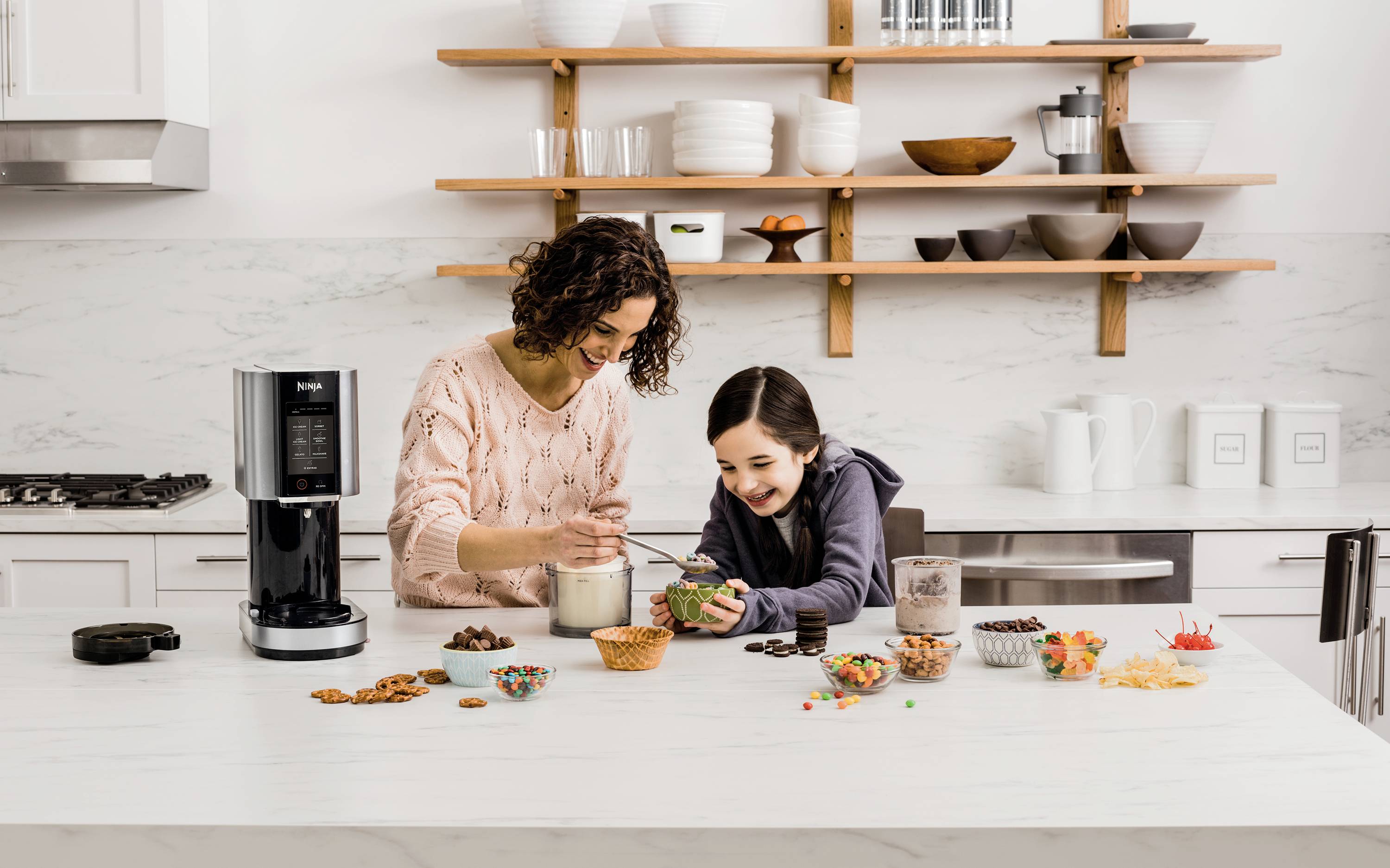 Eine Frau und ein Kind dekorieren freudig Cupcakes in einer modernen Küche. Sie verwenden verschiedene Toppings, die auf dem Tisch verteilt sind.