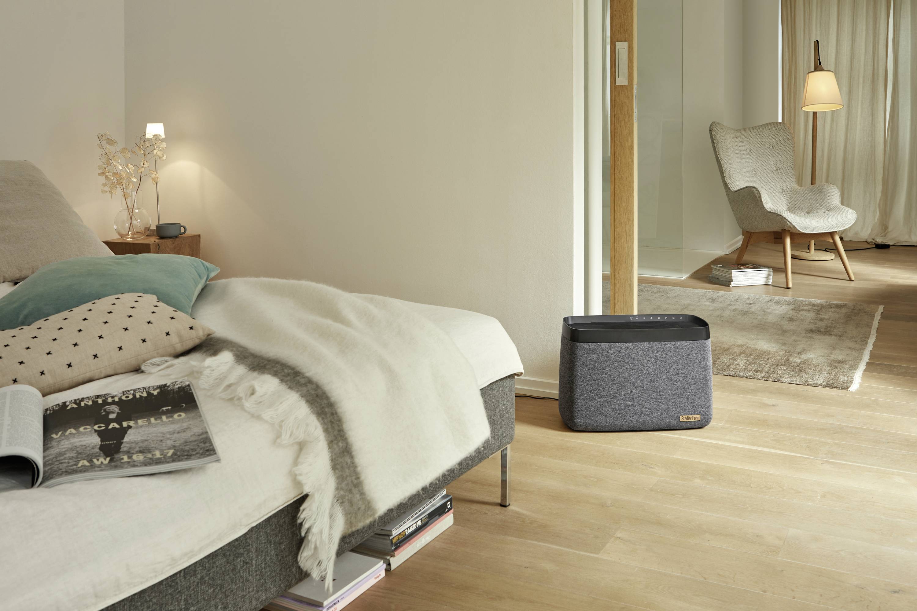 Ein modern eingerichtetes Schlafzimmer mit einem grauen Lautsprecher auf dem Boden, links ein Bett mit Kissen und Decken, rechts ein Sessel.