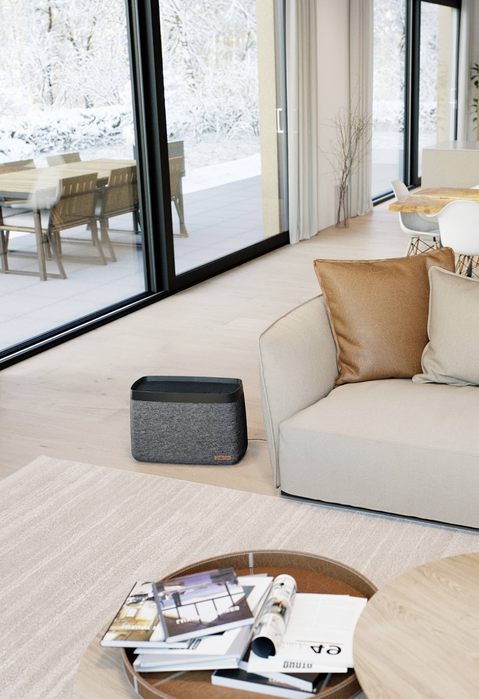 Modernes Wohnzimmer mit großen Fenstern, die auf eine verschneite Terrasse blicken. Ein grauer Lautsprecher steht neben einem beigen Sofa.