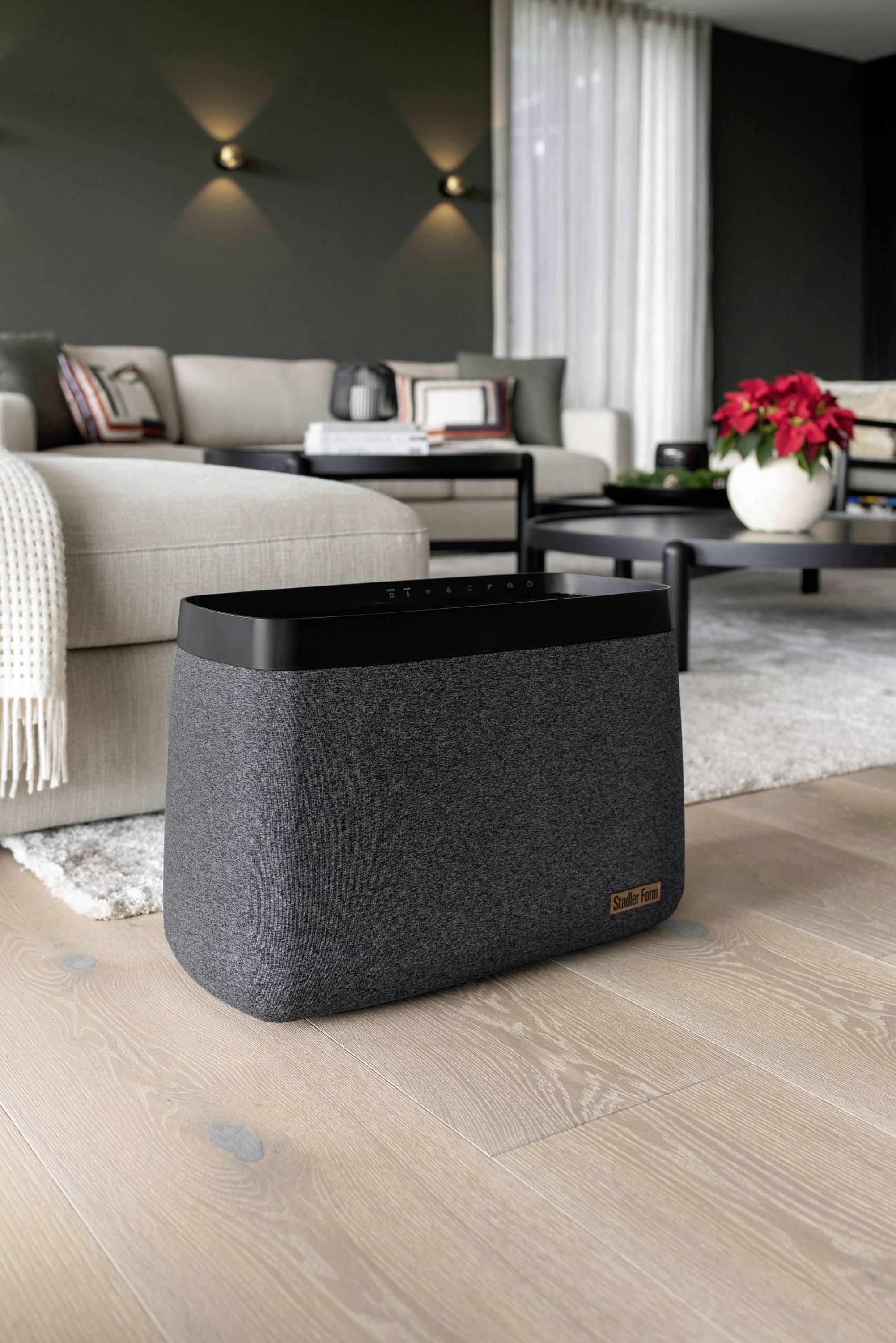 Ein grauer Lautsprecher steht auf einem Holzboden in einem modernen Wohnzimmer. Im Hintergrund ein beiges Sofa und ein Tisch mit Blumen.