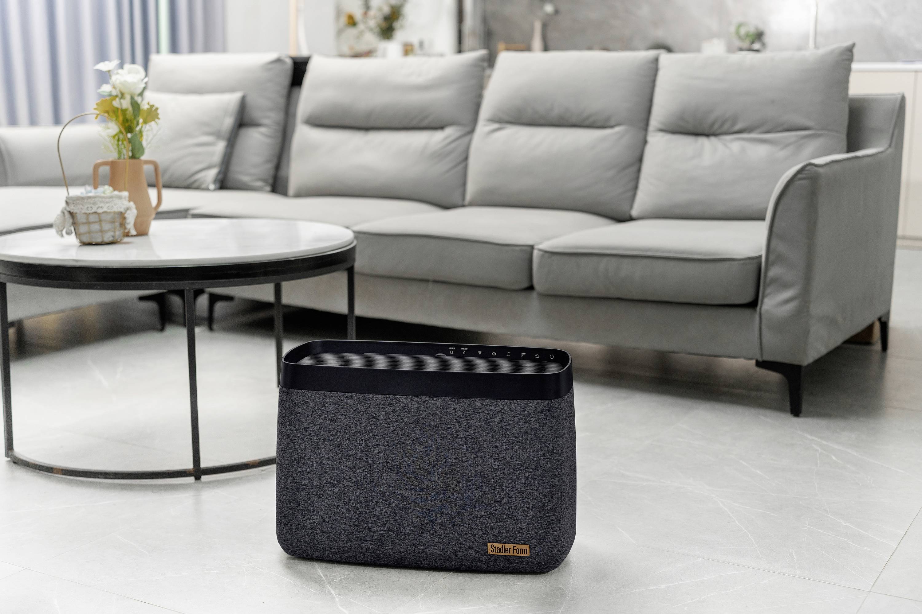 Ein grauer Luftreiniger steht in einem modernen Wohnzimmer mit einem grauen Sofa und einem runden Couchtisch im Hintergrund.