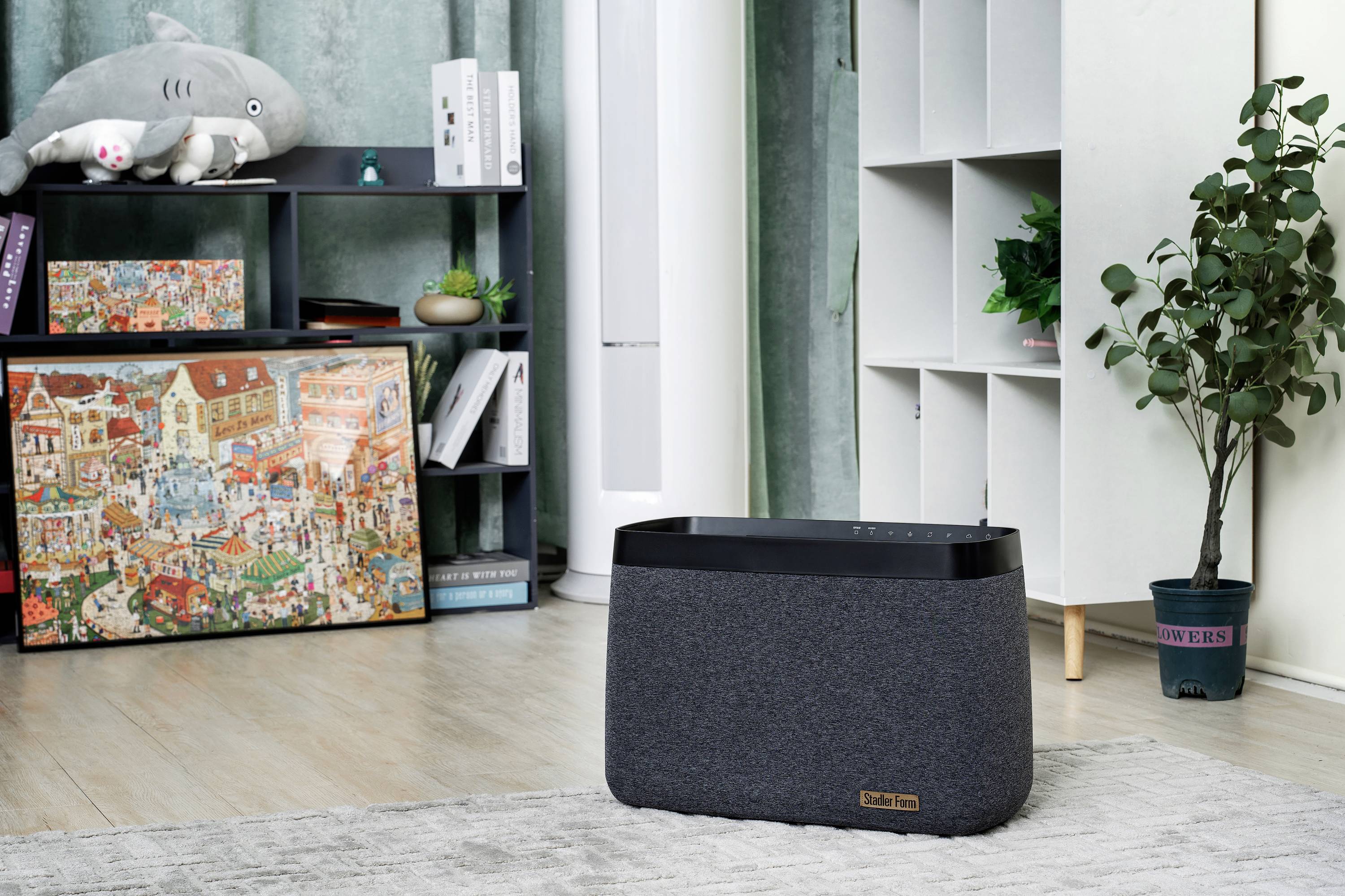 Ein Wohnzimmer mit einem grauen Luftreiniger auf dem Boden, einem Regal mit Pflanzen und einem bunten Puzzle-Leinwandbild an der Wand.