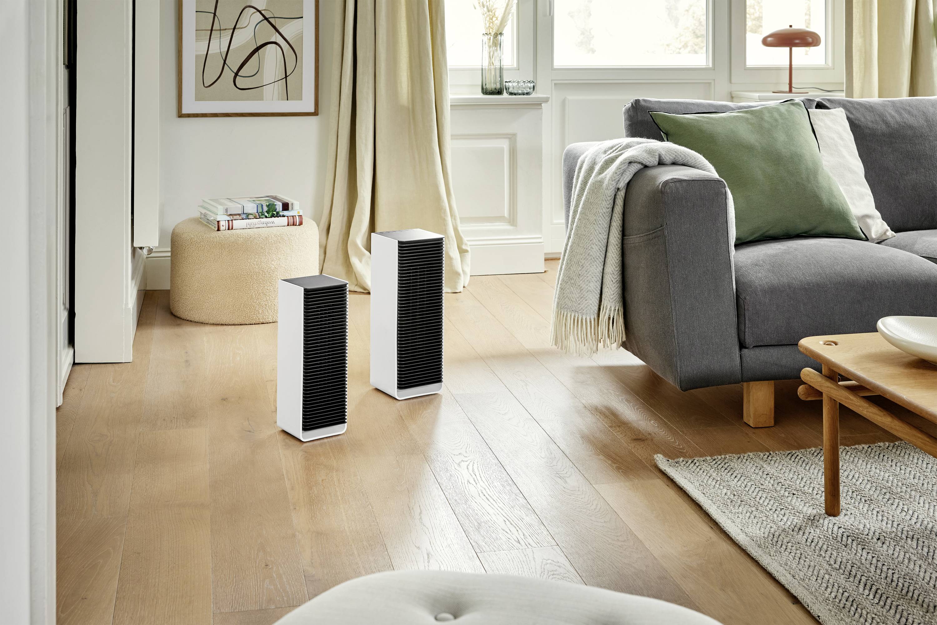 Zwei schwarze Lautsprecher stehen auf einem Holzboden in einem Wohnzimmer mit einem grauen Sofa und einem grünen Kissen.