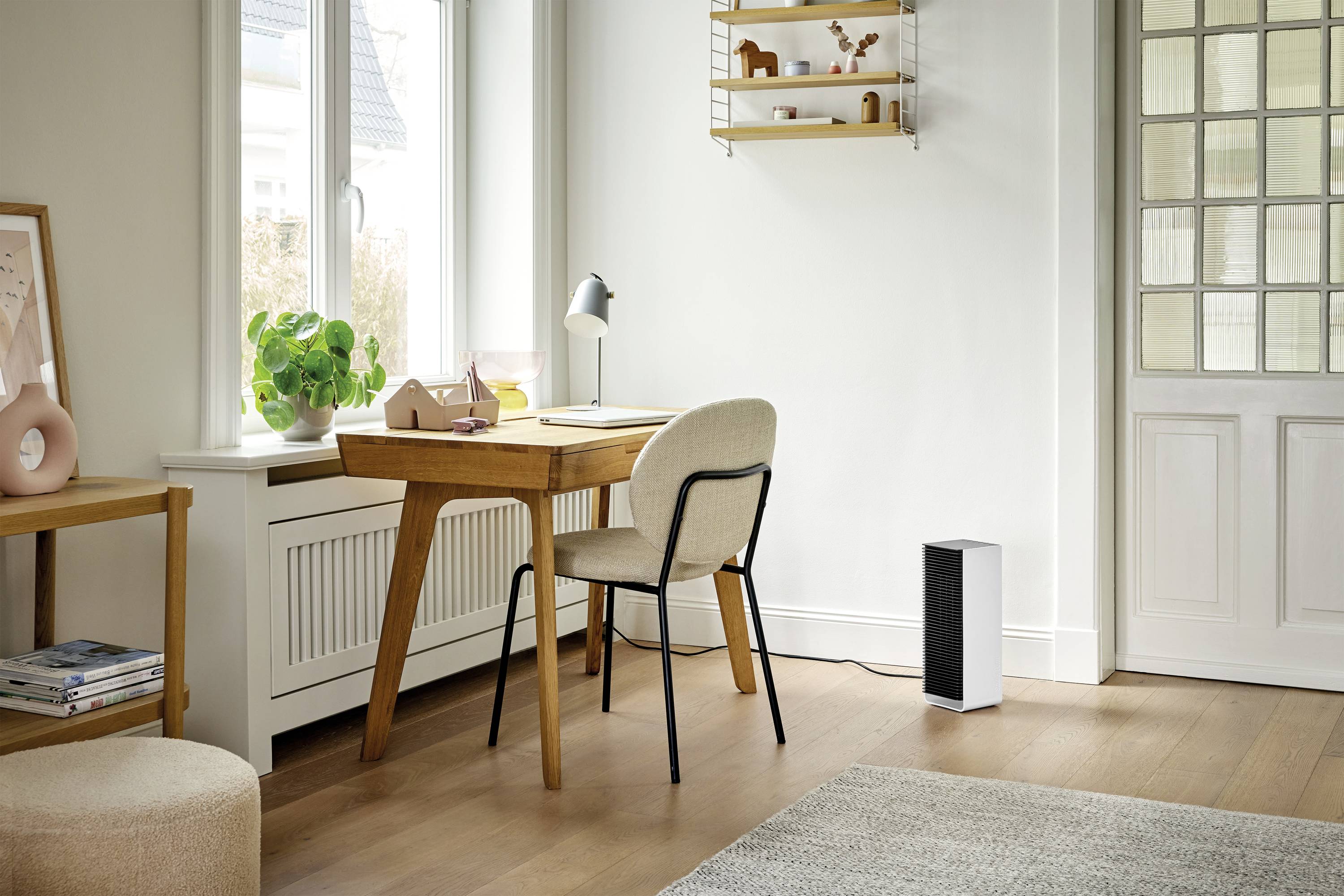 Helles, minimalistisches Arbeitszimmer mit Holzschreibtisch, beige Stuhl, Pflanzen, Tischlampe und Lufreiniger vor einem Fenster.