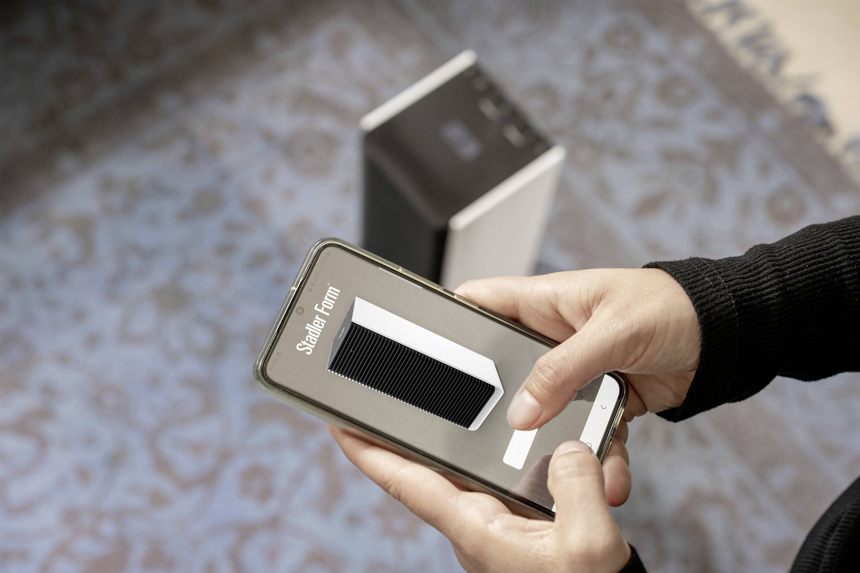 Eine Person hält ein Smartphone, auf dem ein Bild eines Lautsprechers zu sehen ist, während sie neben einem ähnlichen Lautsprecher steht.