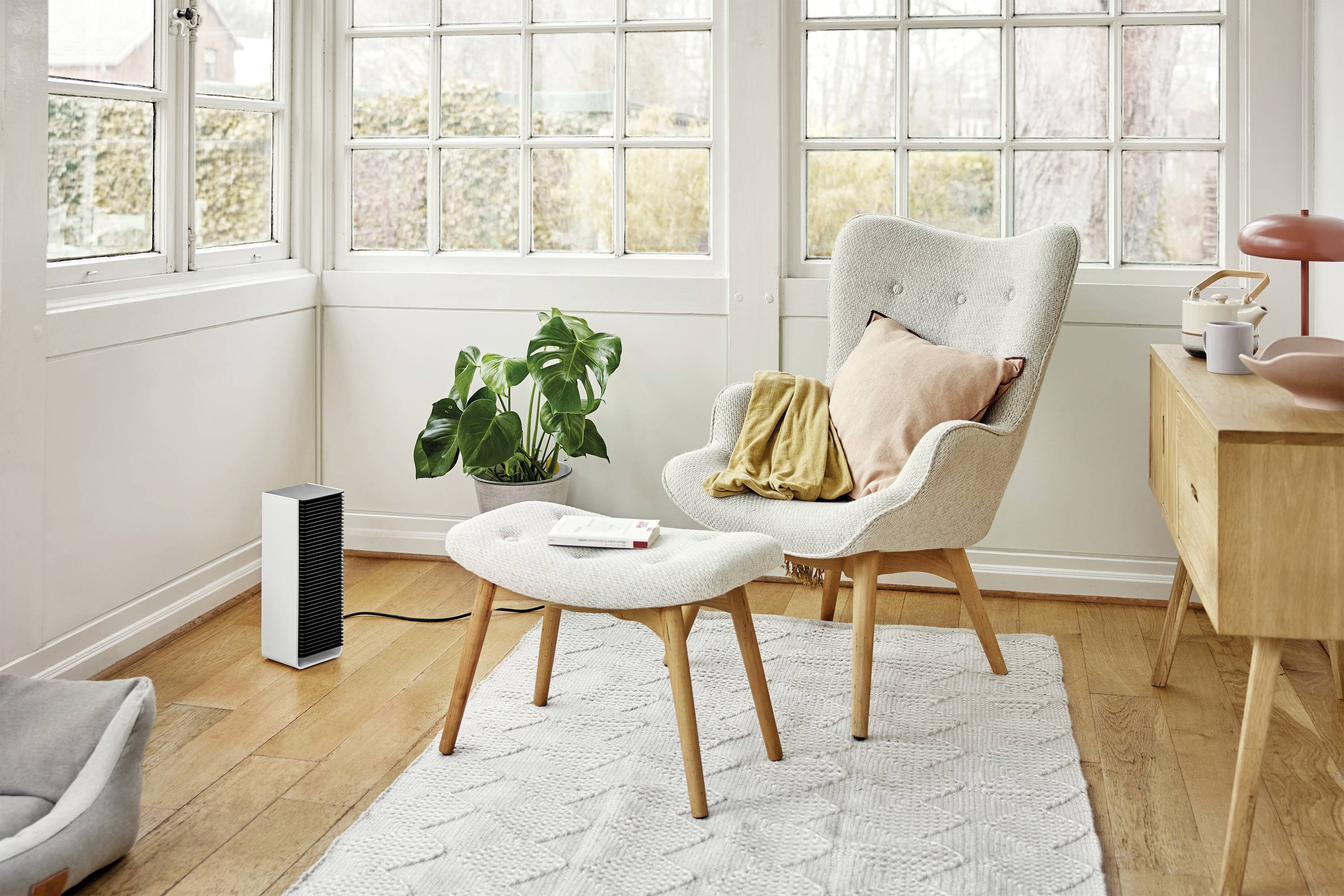 Ein gemütliches Wohnzimmer mit einem weißen Sessel, einem passenden Fußhocker, einer Pflanze und einem kleinen schwarzen Lautsprecher auf einem Teppich.