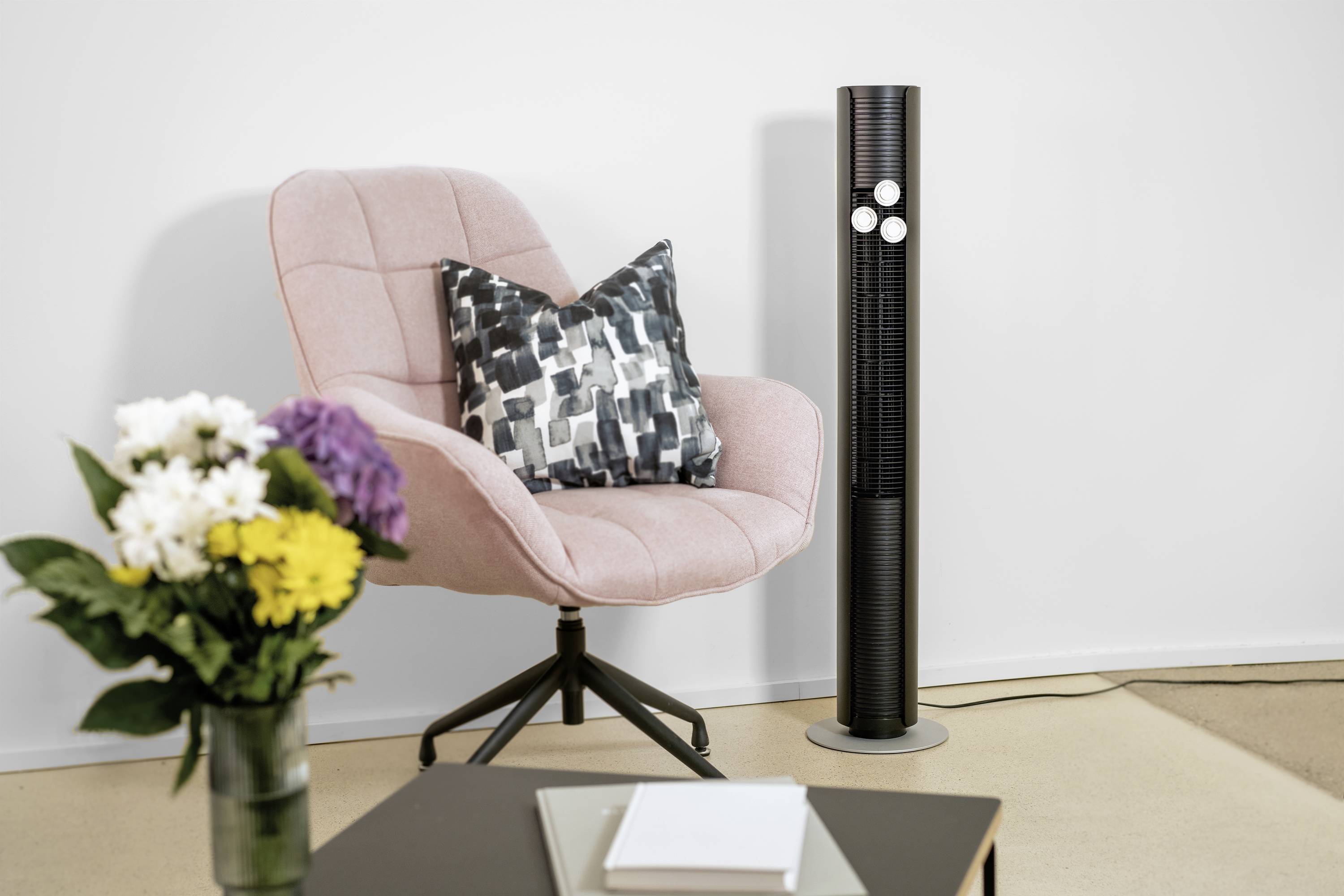 Ein rosa Sessel mit gemustertem Kissen neben einem schwarzen Turmventilator. Im Vordergrund ein Tisch mit Bücherstapel, Blumen in Vase.