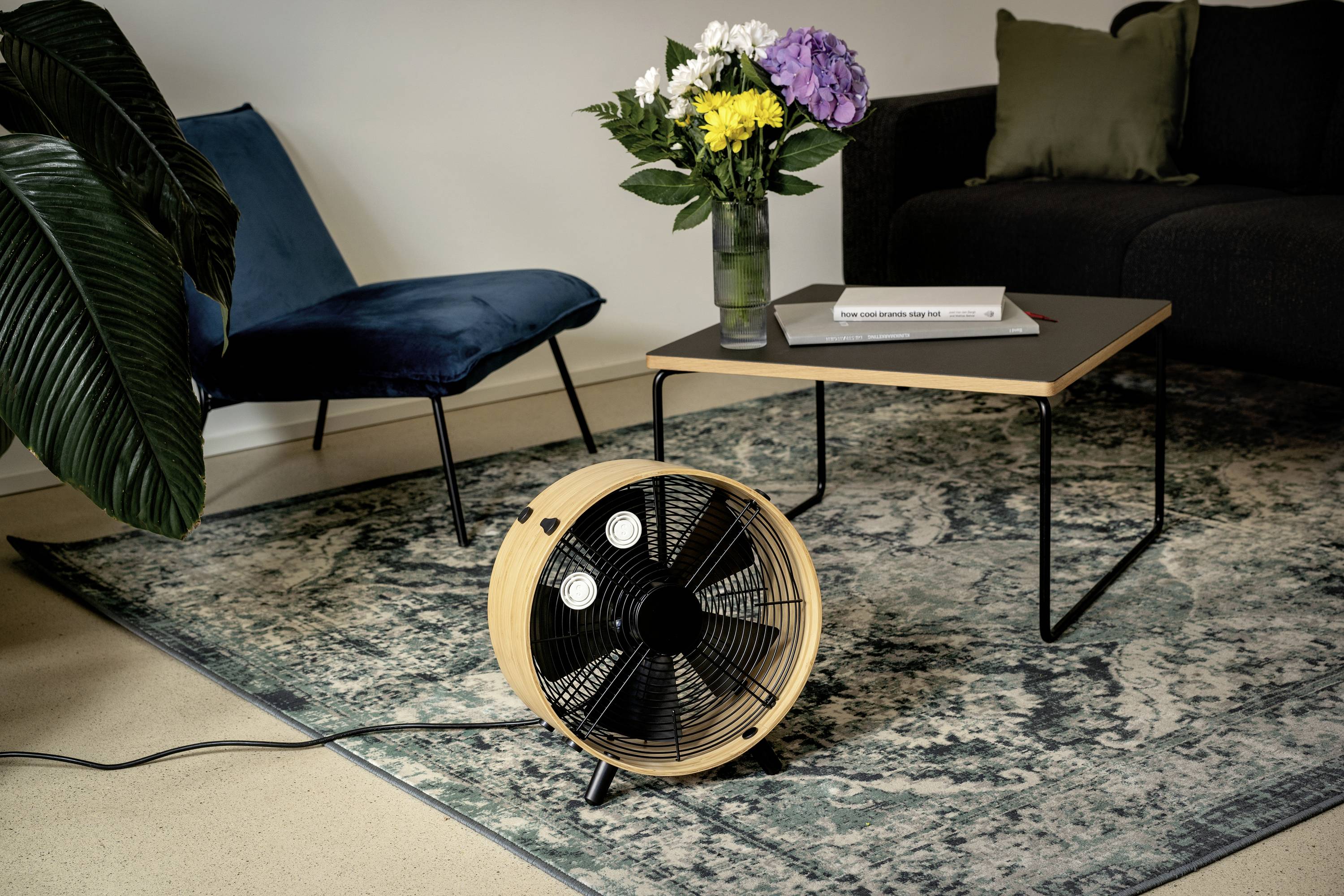 Ein Ventilator auf einem Teppich in einem Wohnzimmer, neben einem Tisch mit Blumen und einem Buch. Im Hintergrund ein Sofa und ein Stuhl.