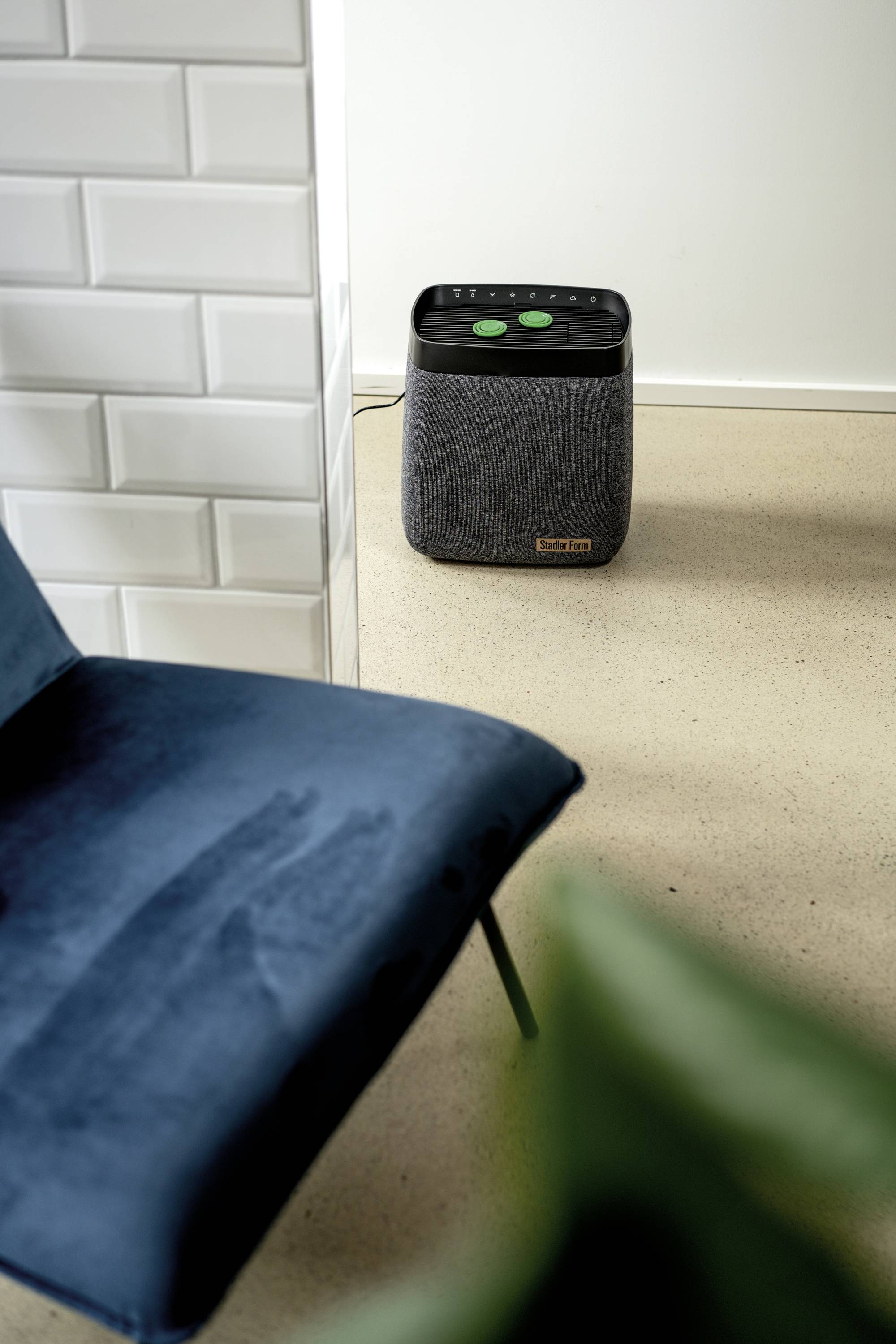 Ein blauer Stuhl steht auf einem beigen Boden vor einer weißen Wand. Im Hintergrund steht ein graues Gerät mit schwarzen und grünen Knöpfen.