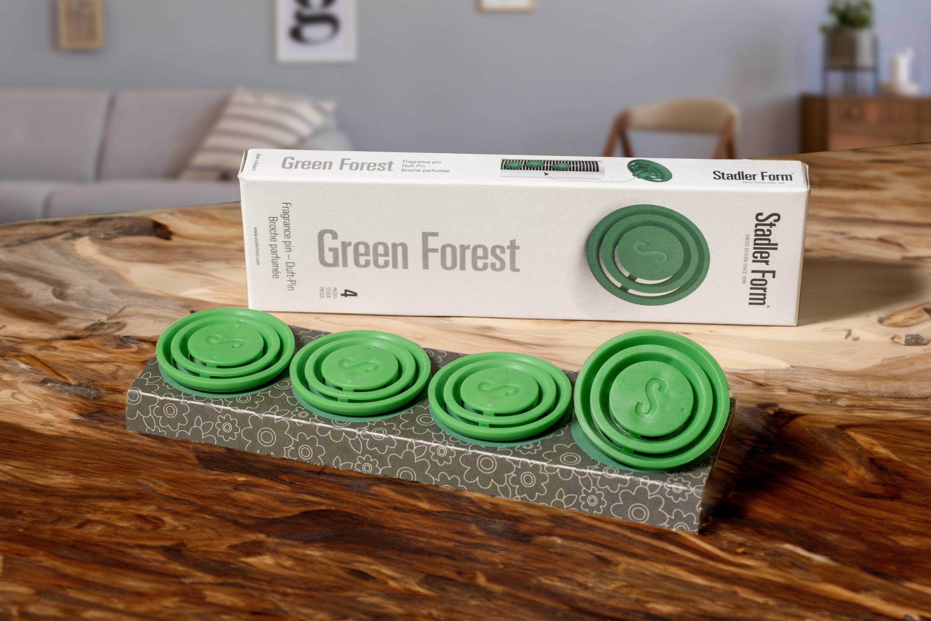 Grüne Duftplättchen auf einem Holztisch. Verpackung mit dem Text 'Green Forest' im Hintergrund. Wohnzimmereinrichtung sichtbar.