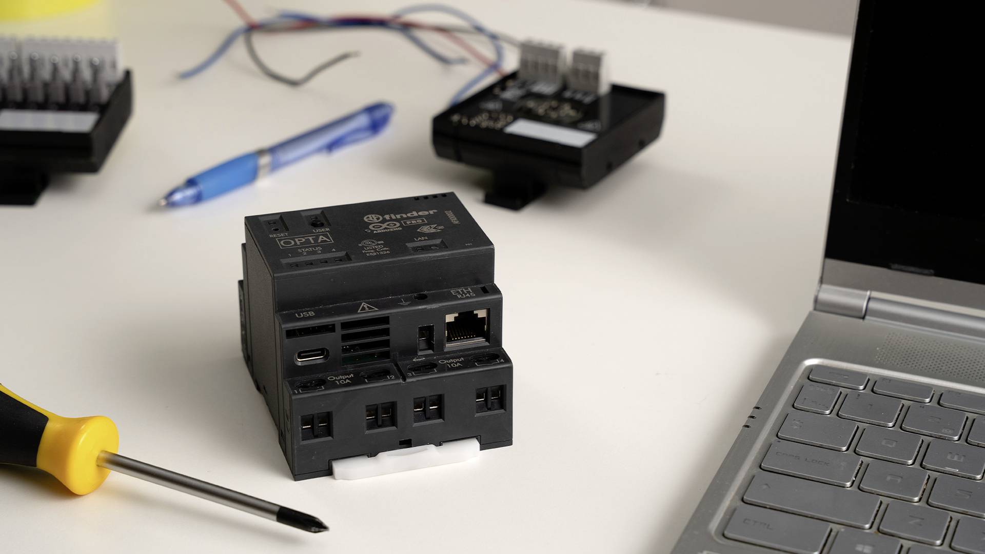 Auf einem Tisch liegt ein schwarzes elektrisches Gerät neben einem Schraubendreher und einem Laptop. Im Hintergrund sind Kabel und ein Stift zu sehen.