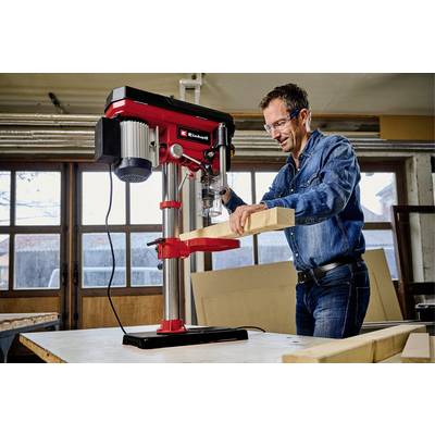 Ein Mann bearbeitet ein Holzstück mit einer roten Standbohrmaschine in einer Werkstatt. Er trägt Schutzbrille und Jeanskleidung.