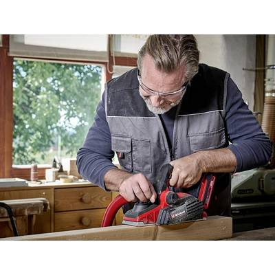 Ein älterer Mann hobelt ein Holzbrett in einer Werkstatt. Er trägt Schutzbrille und Arbeitskleidung. Im Hintergrund ist ein Fenster mit Blick auf Bäume.