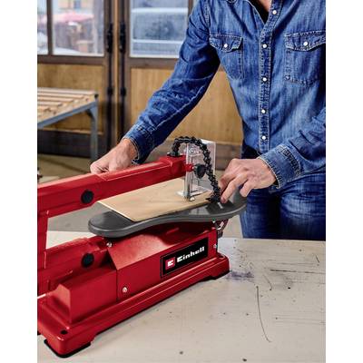 Eine Person in Jeanshemd bedient eine rote Dekupiersäge von Einhell, um ein Holzstück zu schneiden. Die Werkstatt hat Holzwände und Fenster.